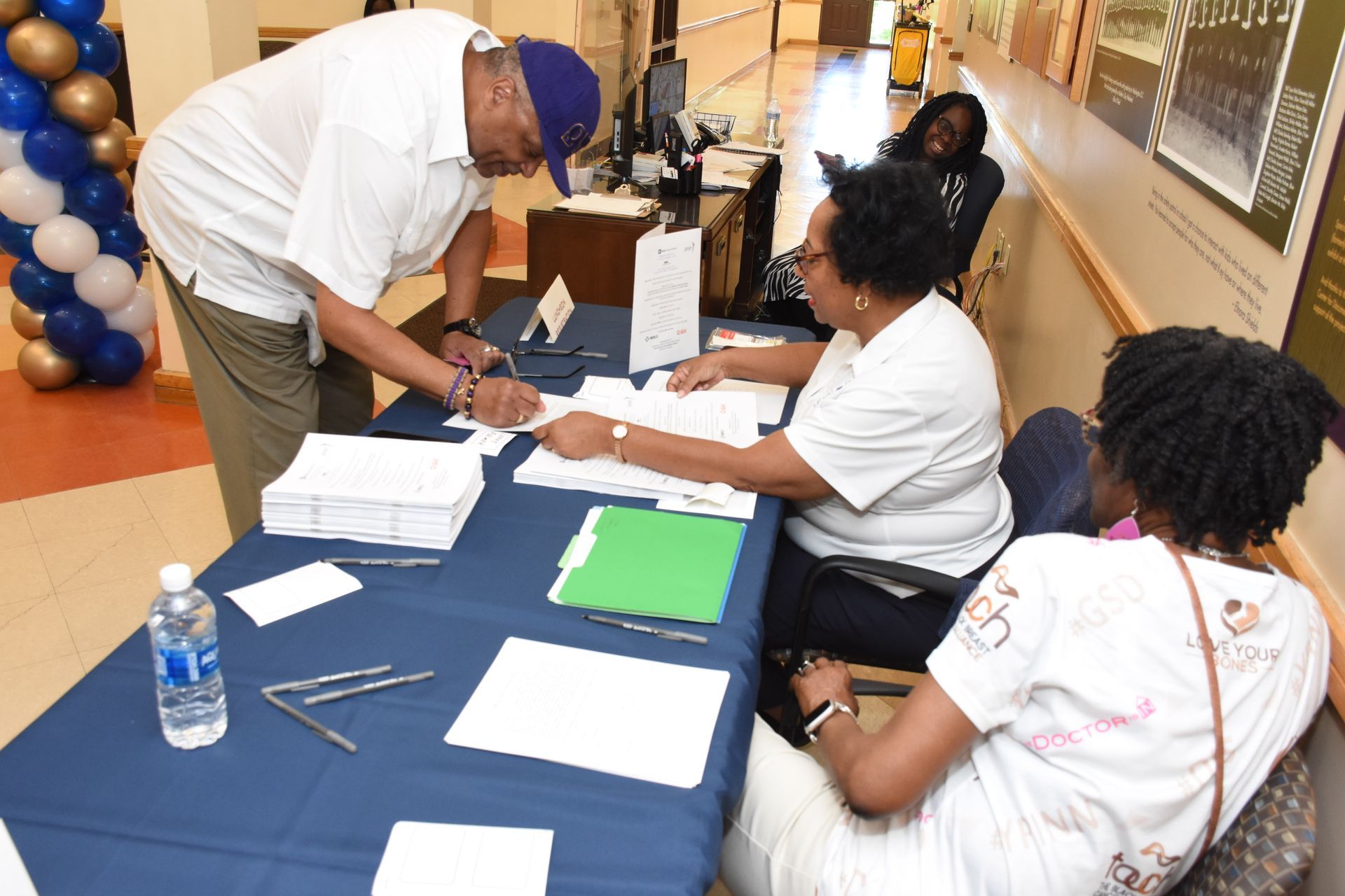 Man signing a form at a table with two women; blue table cloth, papers, water bottle, and balloons.