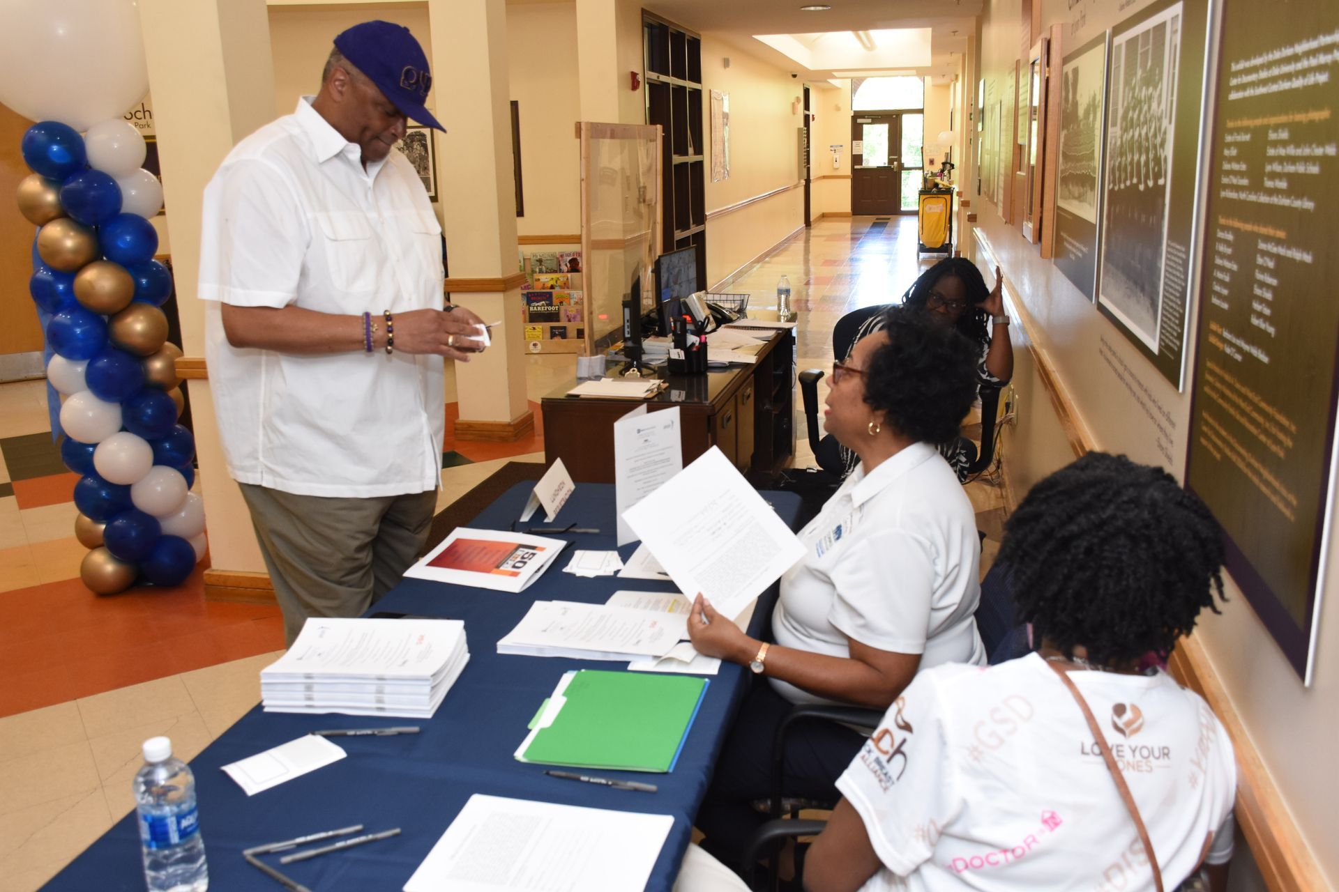 Man at a table with two people. Hallway with balloons. People are looking at paperwork.