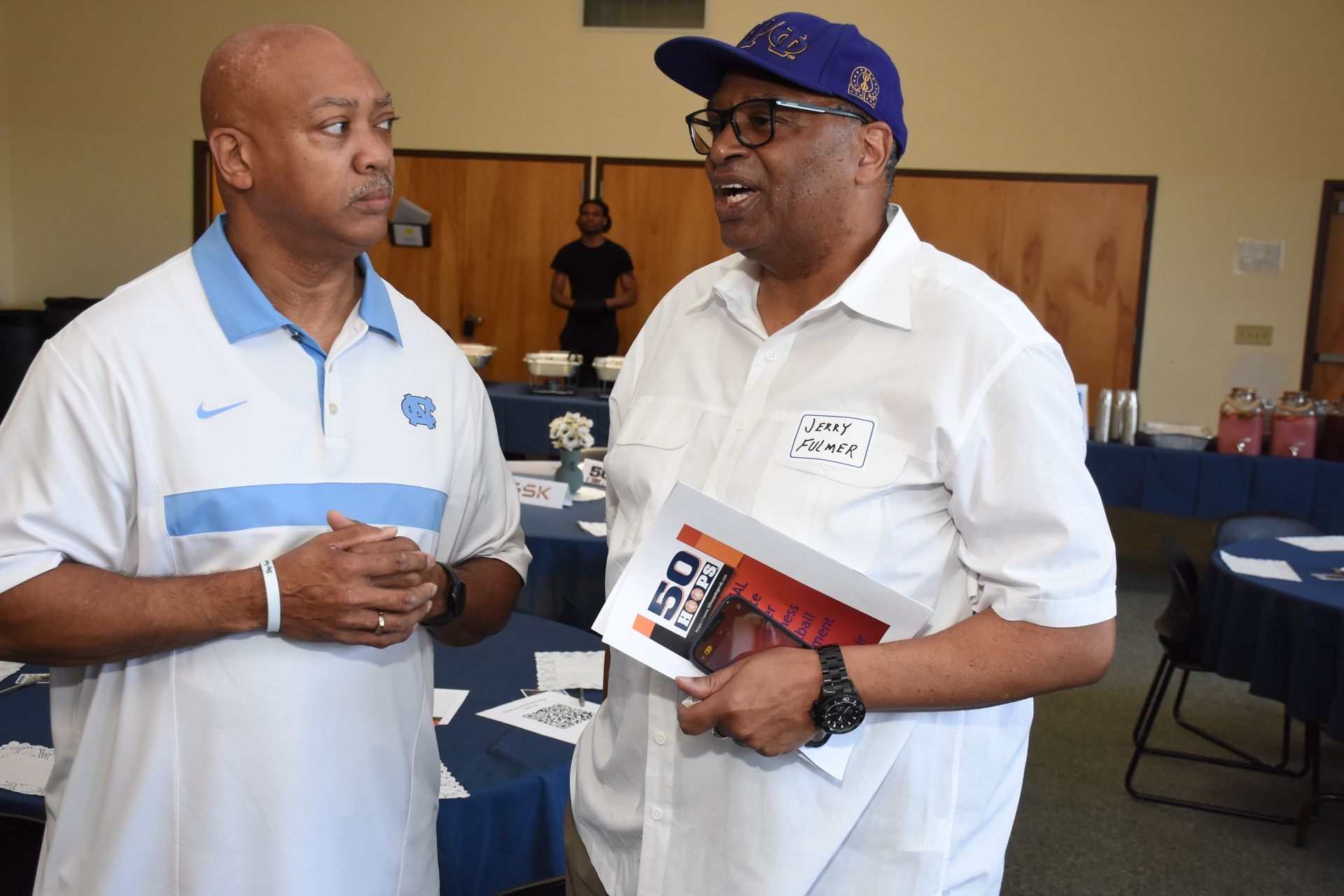 Two men converse, one in a light blue polo, the other in a white shirt and purple cap, at an event.