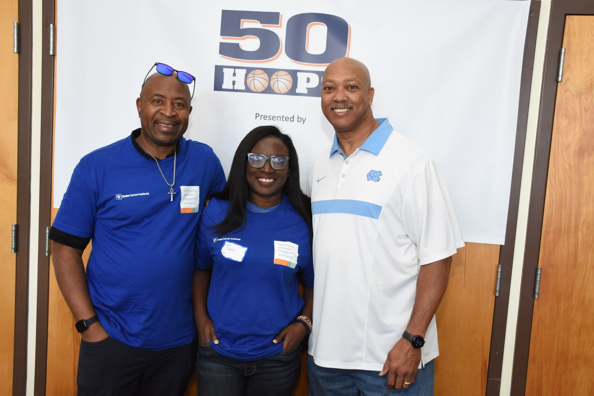 Three people pose for a photo in front of a banner. They wear blue shirts. One man wears sunglasses.