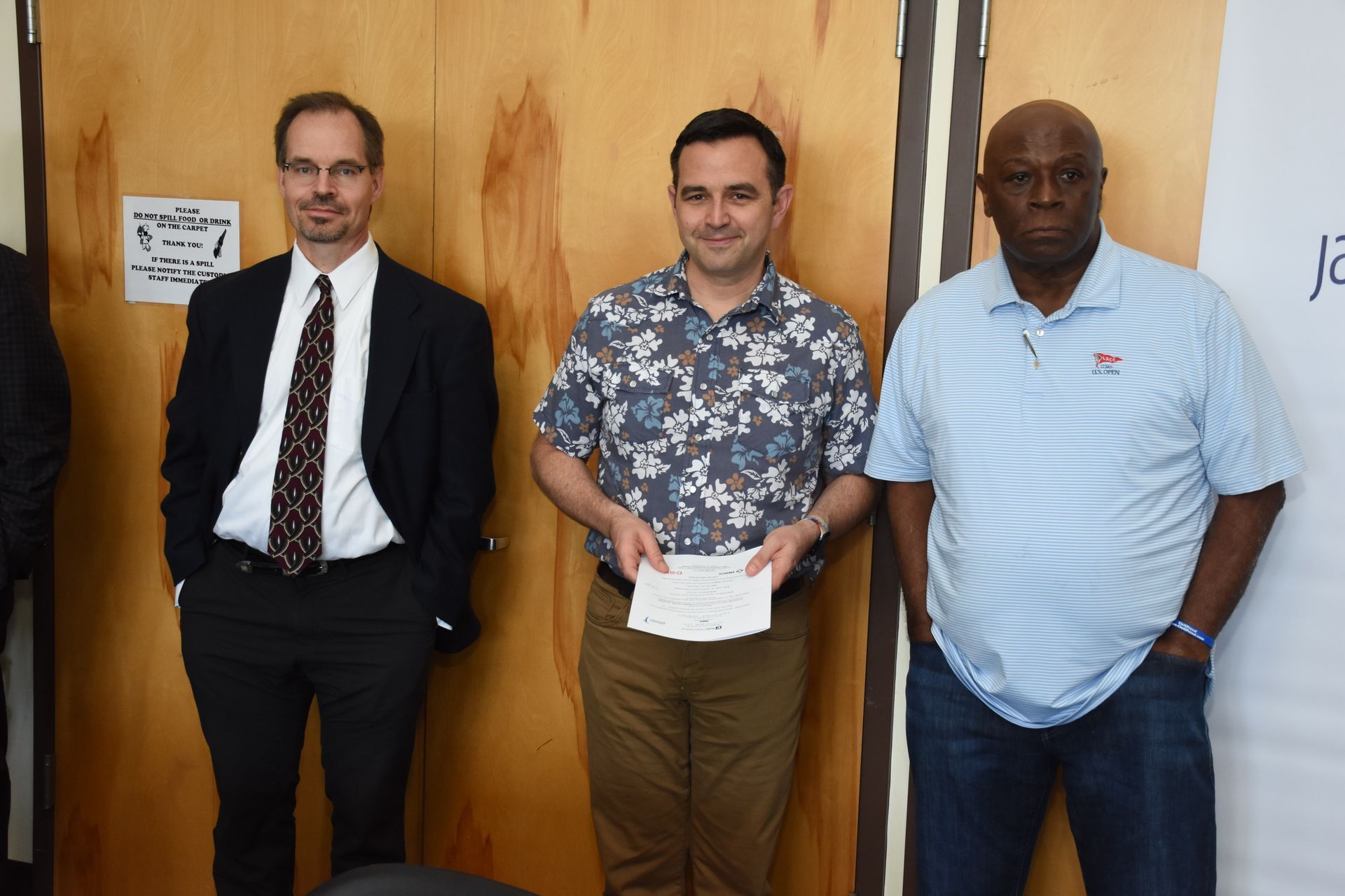 Three men standing, one holding a check.  They are in a room with a wooden wall.