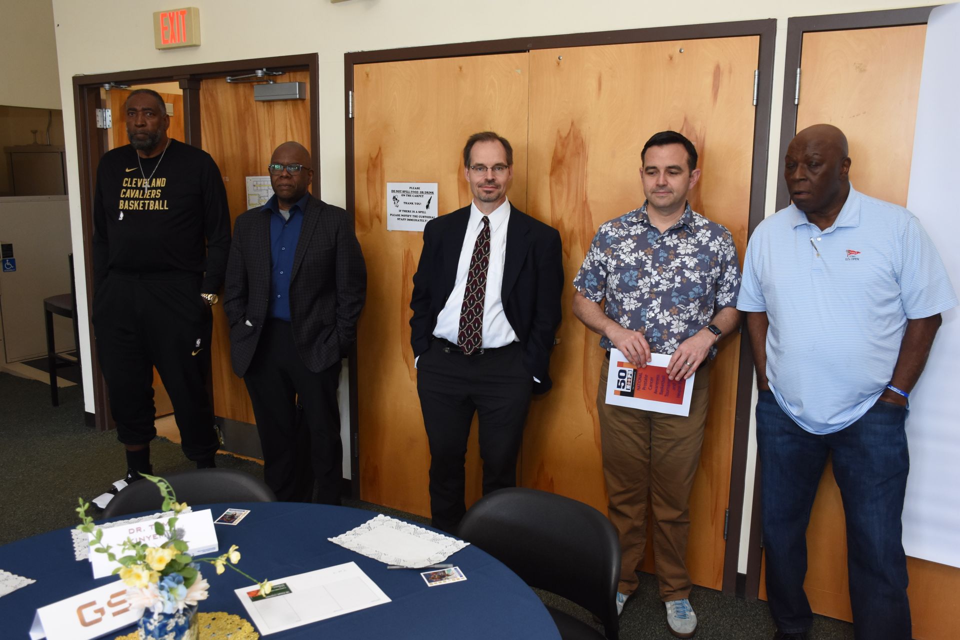 Five men stand in front of wooden doors. One holds papers, others in suits or casual wear, near a table with flowers.