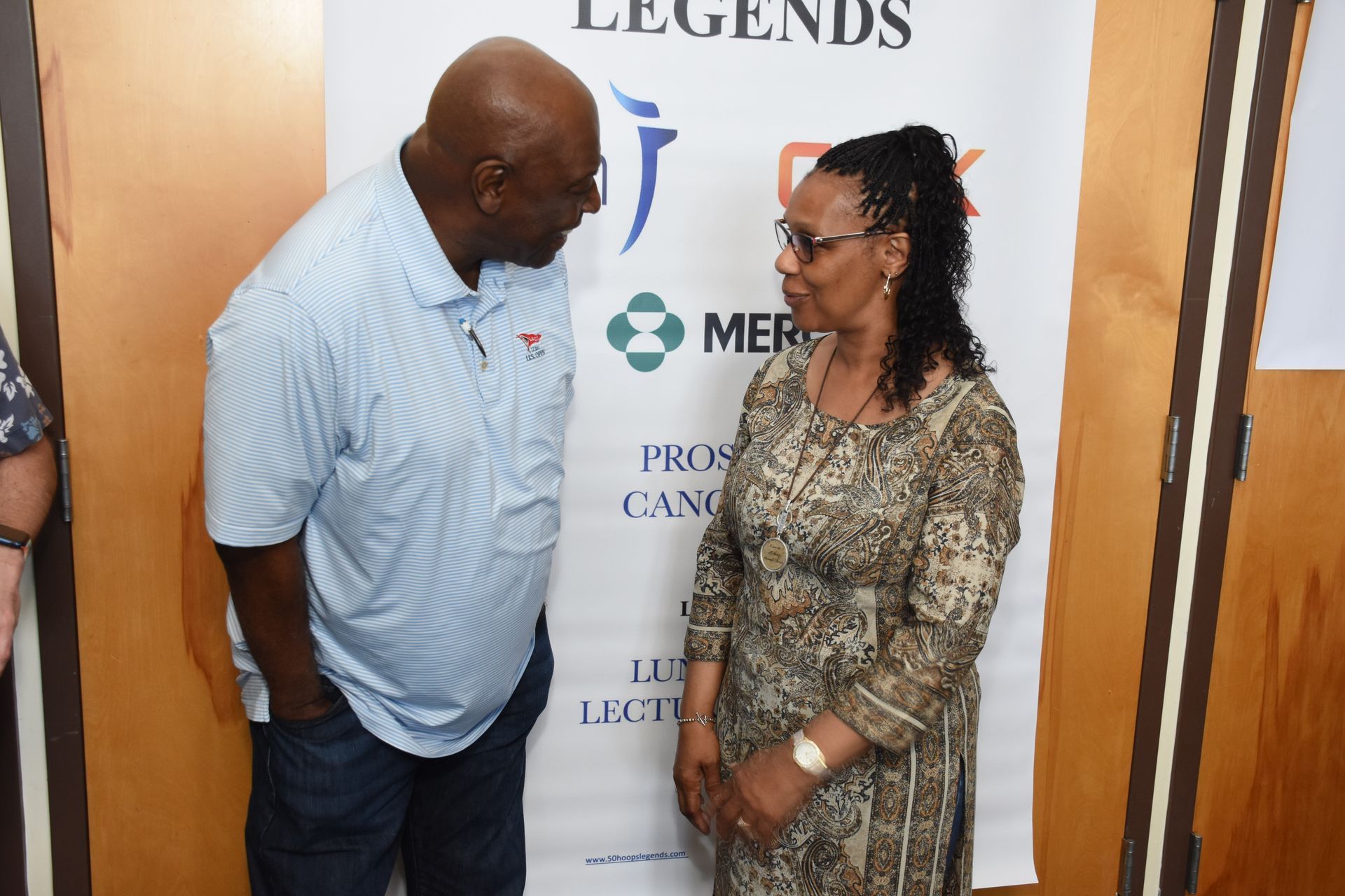Man in blue shirt and woman in patterned dress stand talking near a banner.