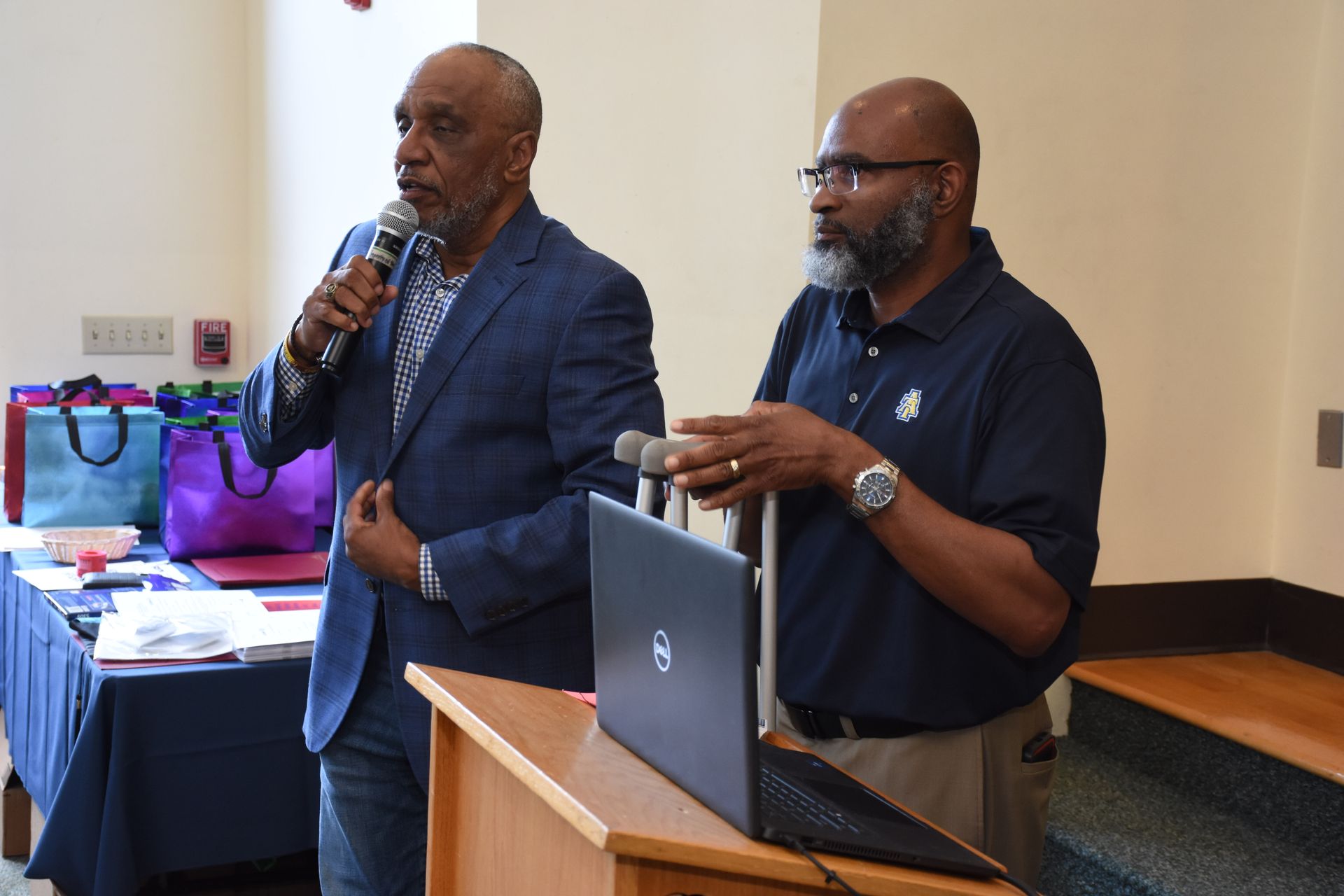 Two men at a podium, one speaking into a microphone. A laptop sits on the podium. Behind them are tables with bags.
