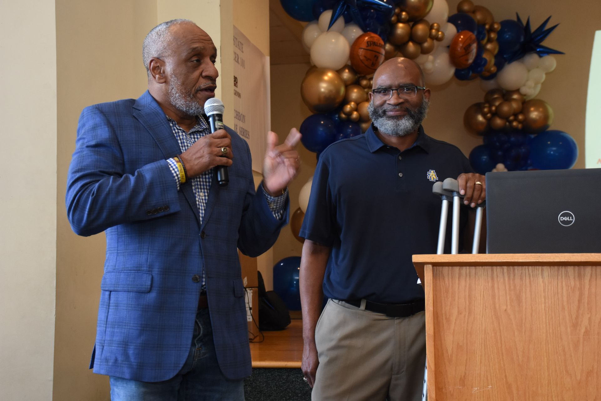 Two men at podium, one speaking with microphone. One uses crutches. Balloons in background.