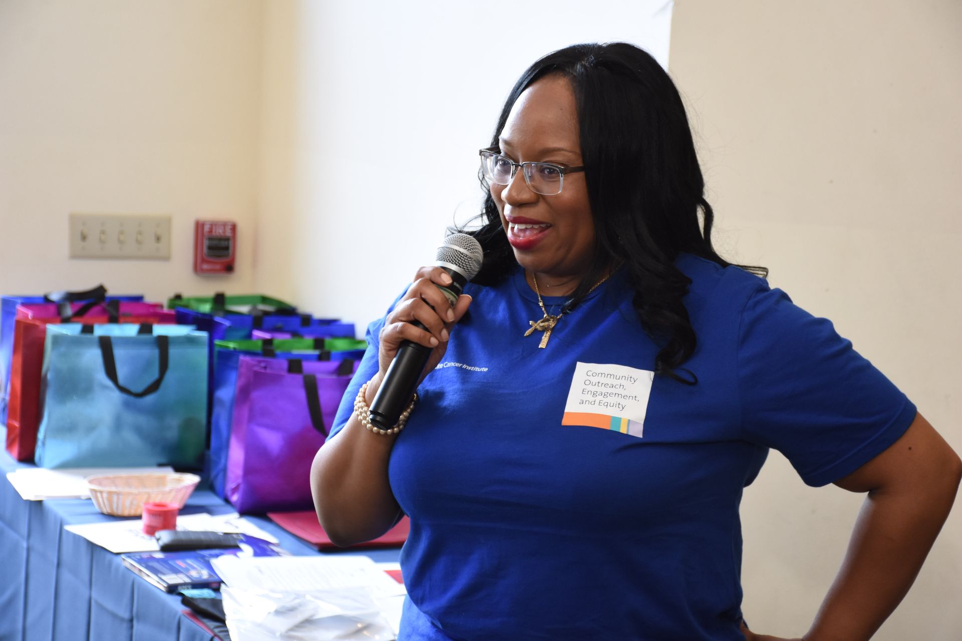 Woman in blue shirt speaking into a microphone with colorful bags on a table in the background.