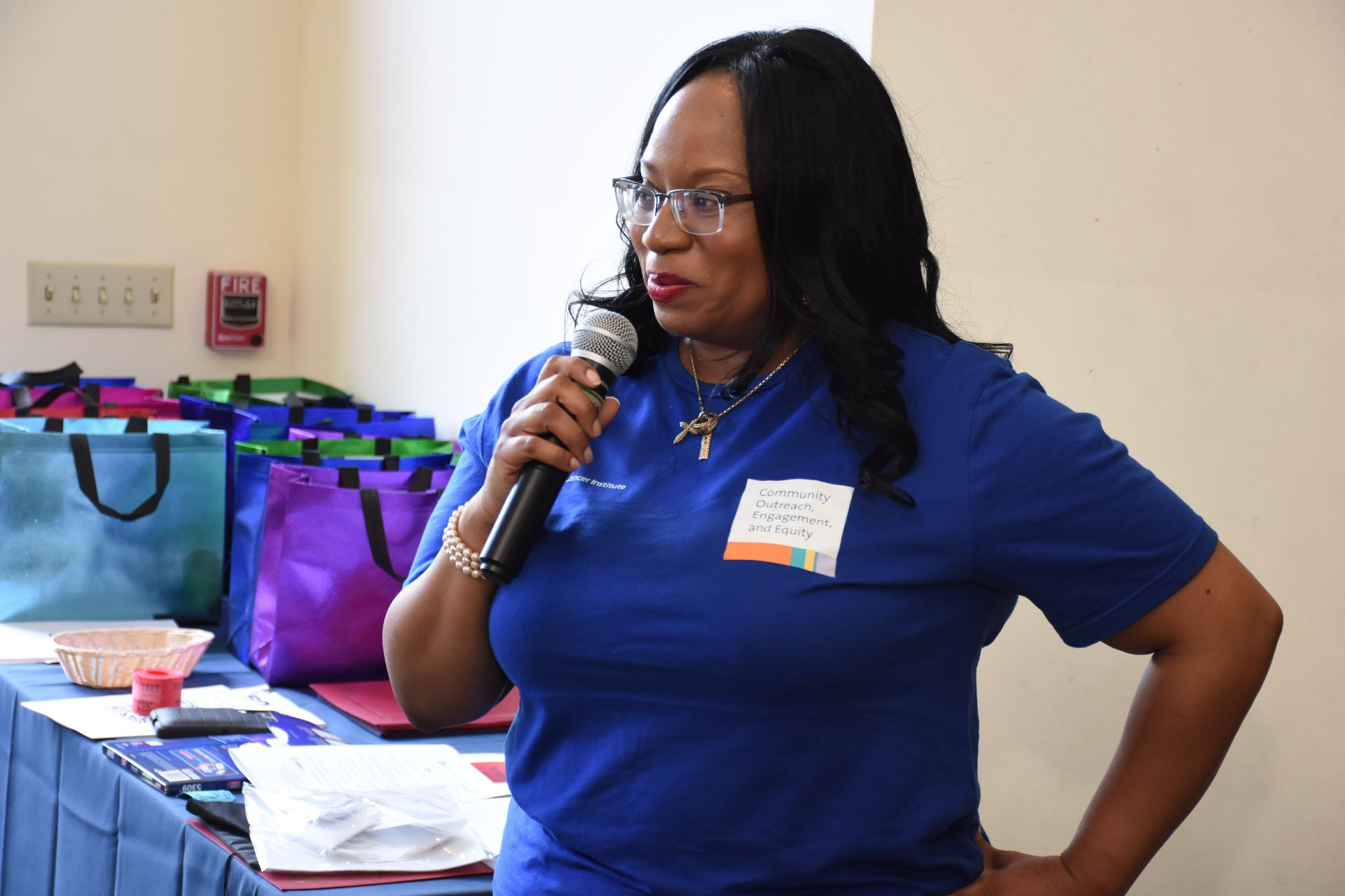 Woman in blue shirt speaking into a microphone. Standing next to a table with bags. Indoors.