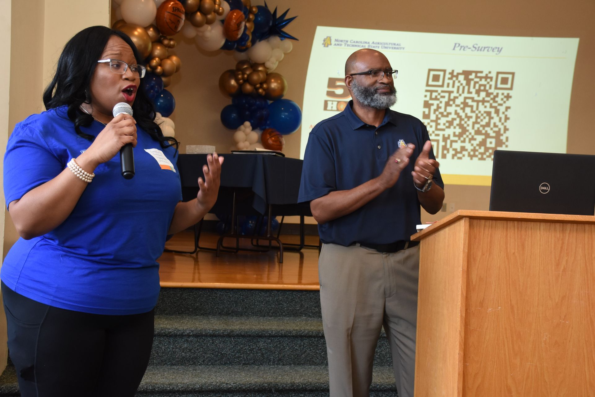Woman speaking into a microphone, man clapping, in front of a presentation screen, balloons, and stairs.