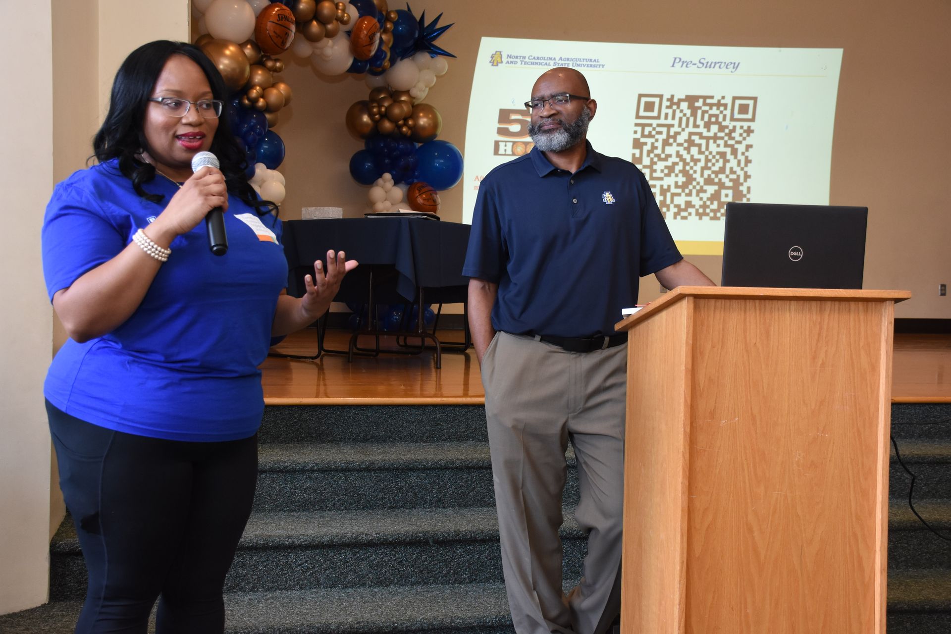 Two people at a podium, one speaking into a microphone. A presentation screen is behind them, with balloons.