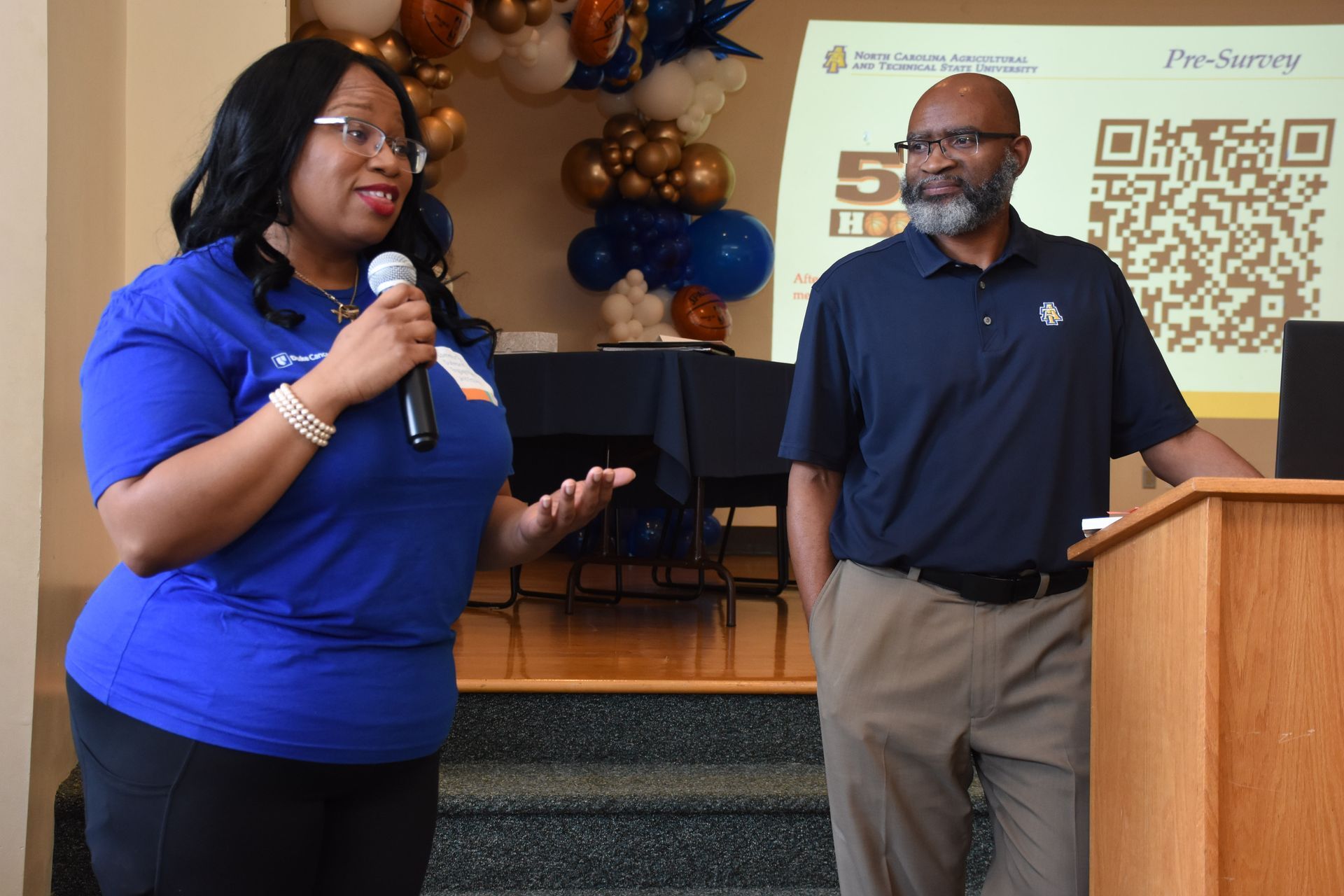 Woman speaking into a microphone, man beside her, both at a podium. Balloons and a presentation screen in the background.