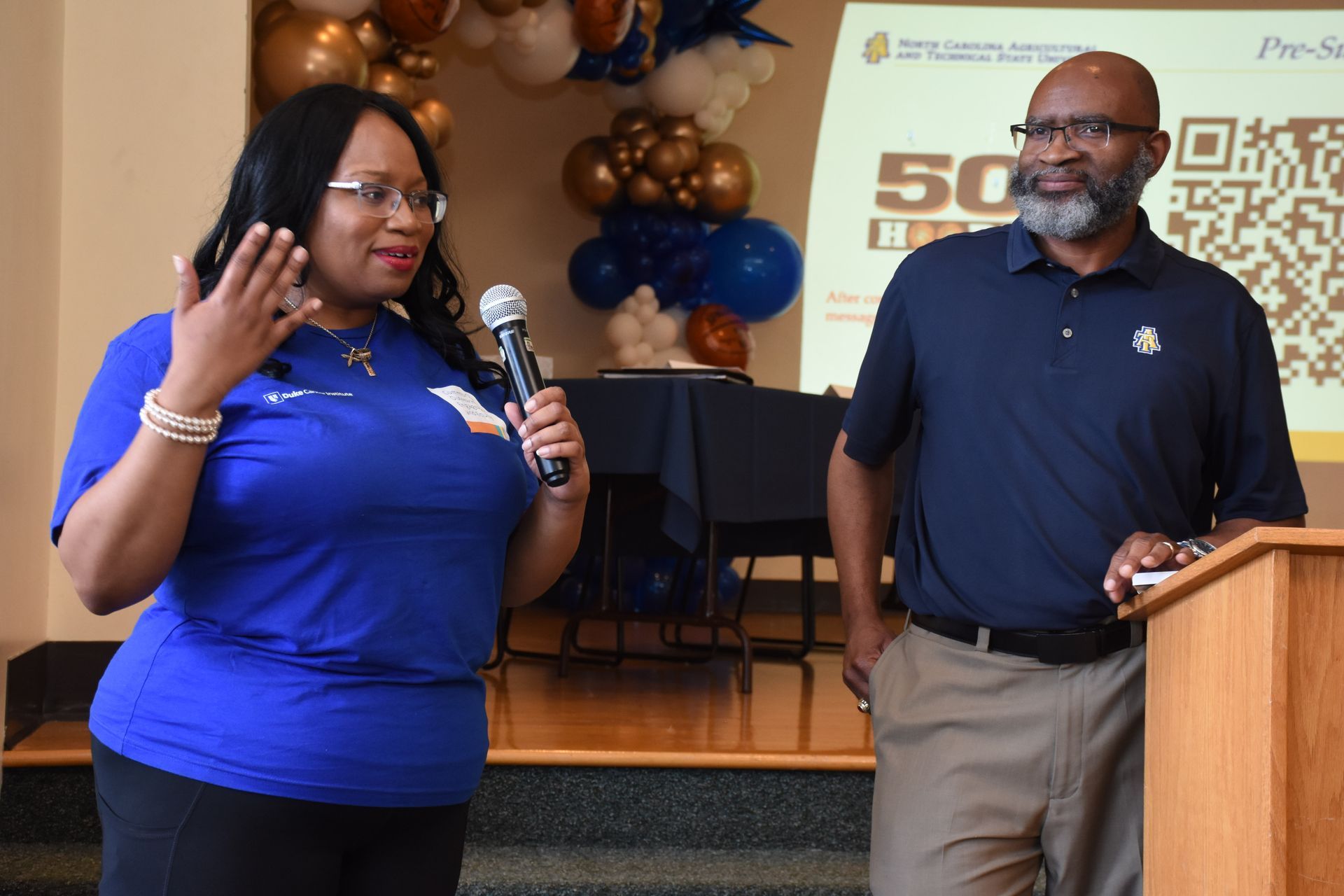 Woman with microphone speaks near a man at a podium, gold and blue balloons in background.