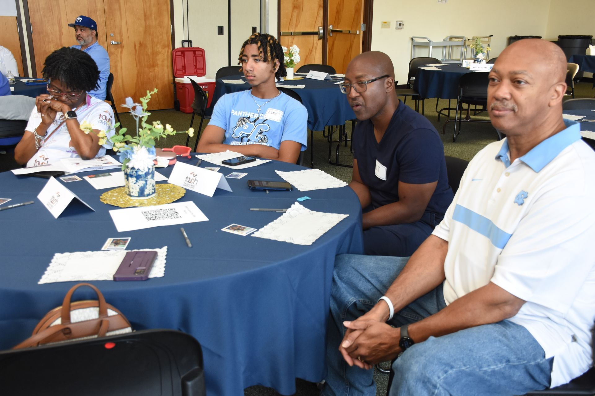 People seated at a table covered with a blue tablecloth, some with name tags.  One man is wearing a light blue polo.