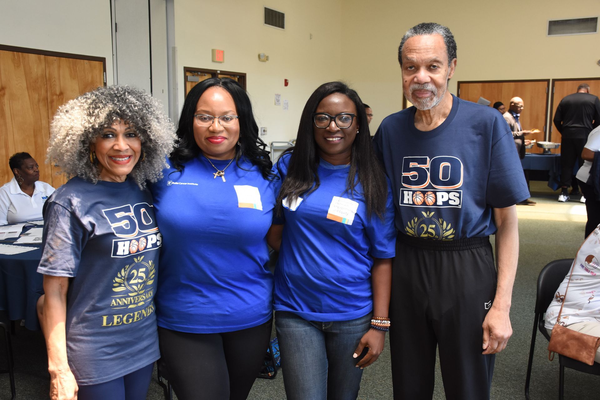 Four people in blue shirts, posing indoors. The shirts read 