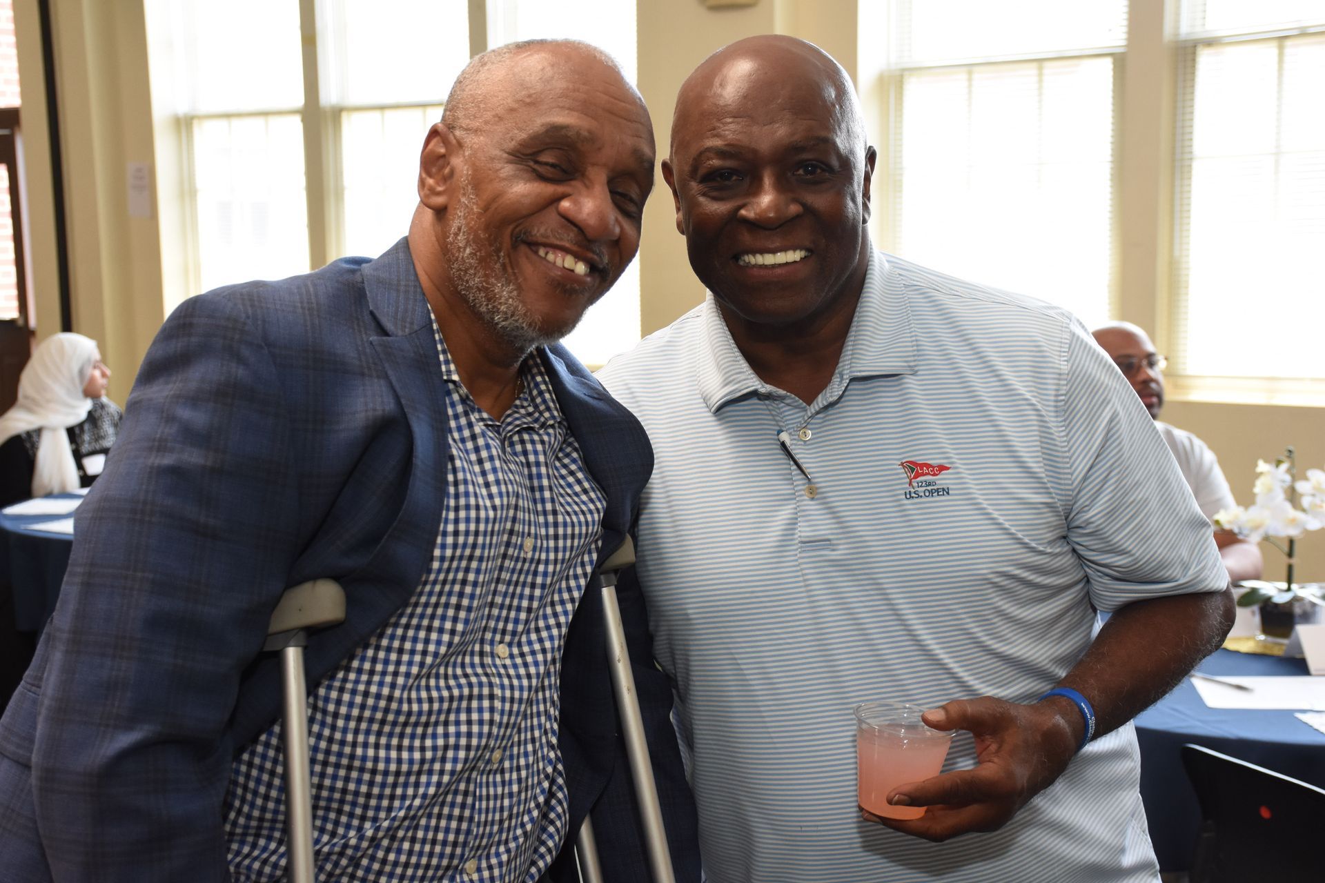 Two men smiling, one with crutches and a blue blazer, the other in a light blue polo. Indoors.