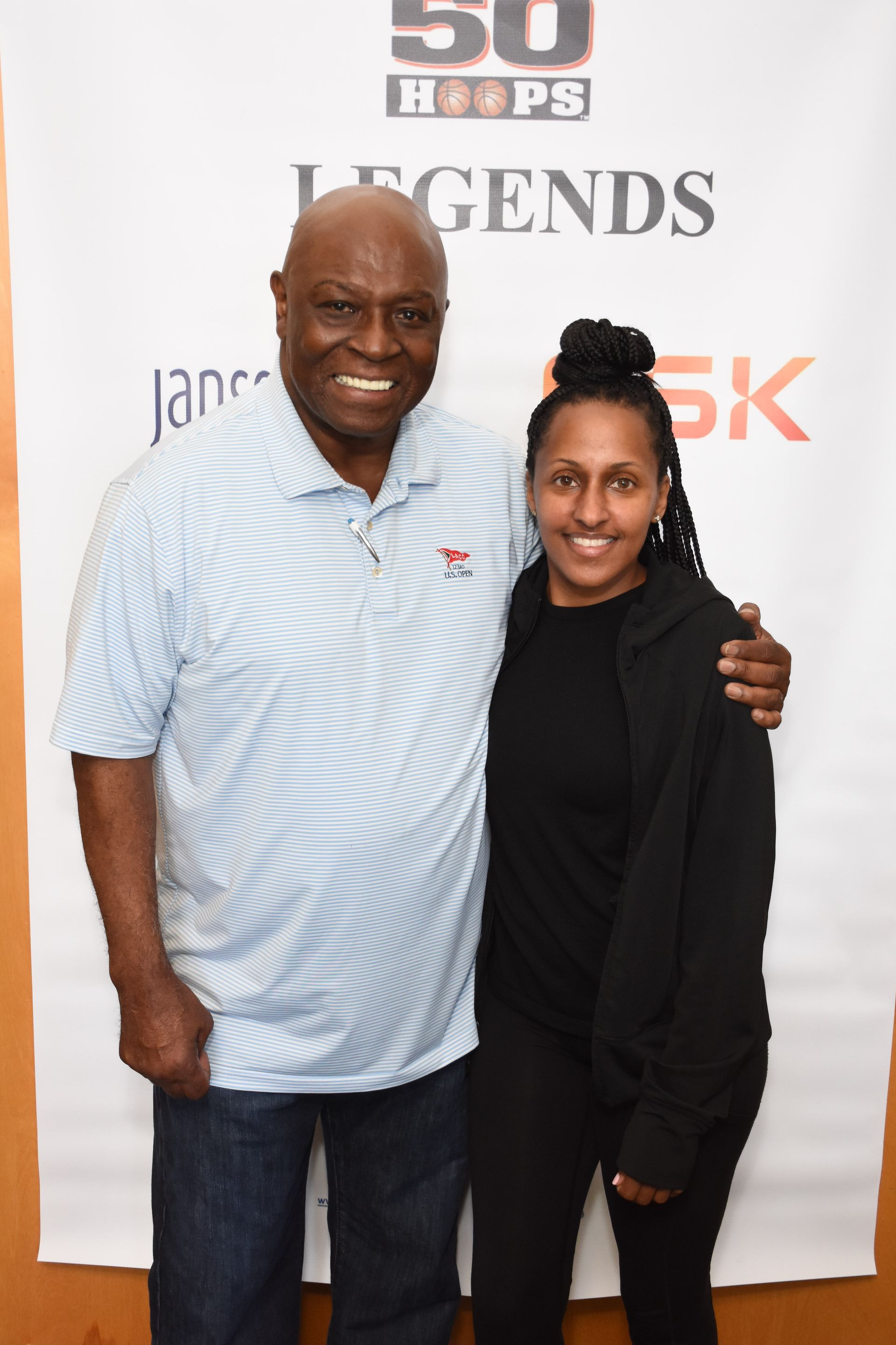 Man in blue shirt and woman in black jacket pose together, smiling, near a banner.
