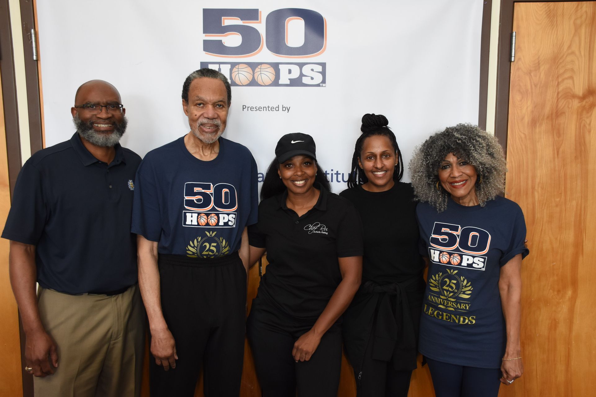 Five people pose in front of a banner. They are wearing “50 Hoops” t-shirts, except for the woman in the center.