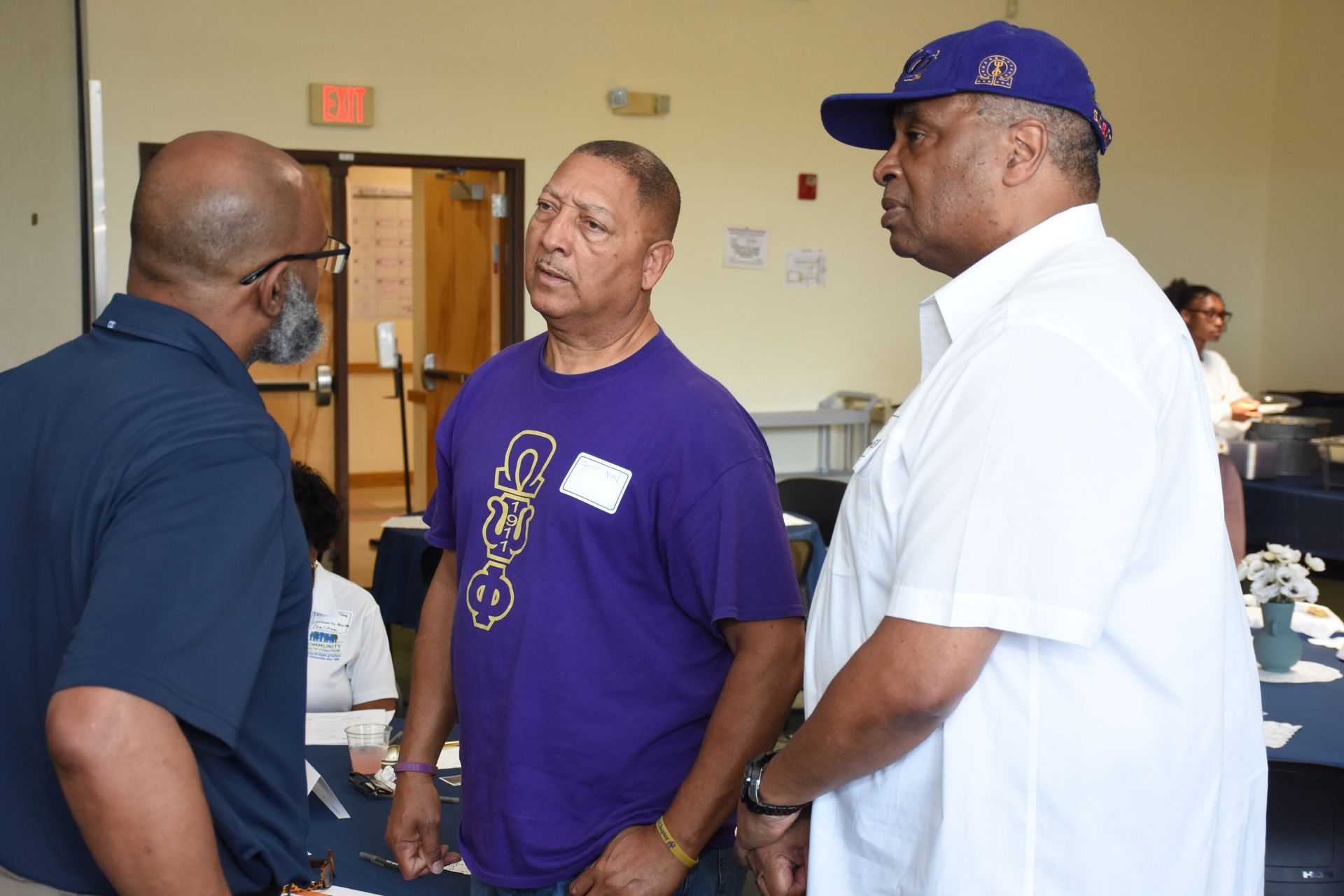 Three men in conversation at an event; two wear purple. A woman can be seen in the background.