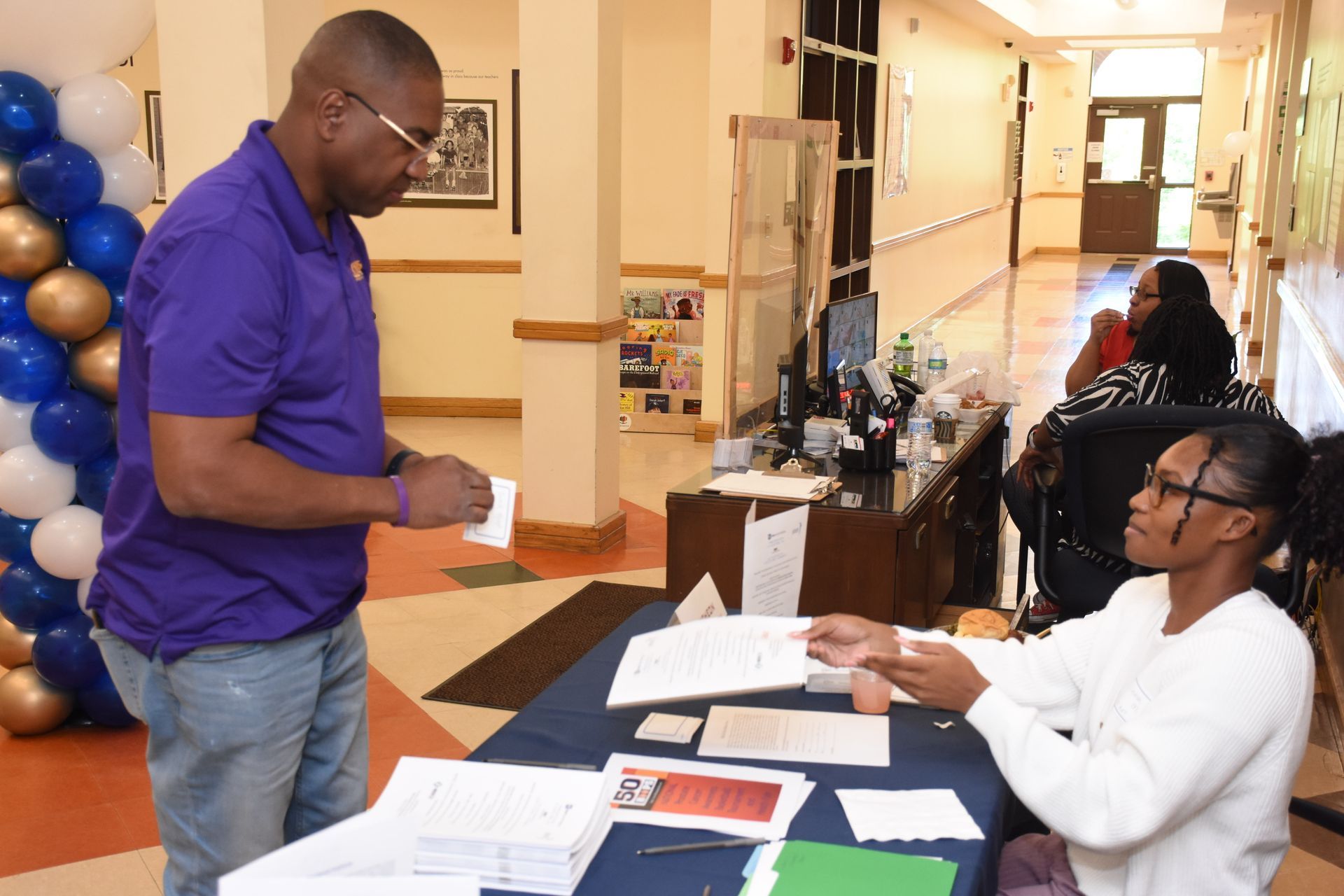 Man in purple shirt at a check-in table. Woman hands him papers. Balloons in background.
