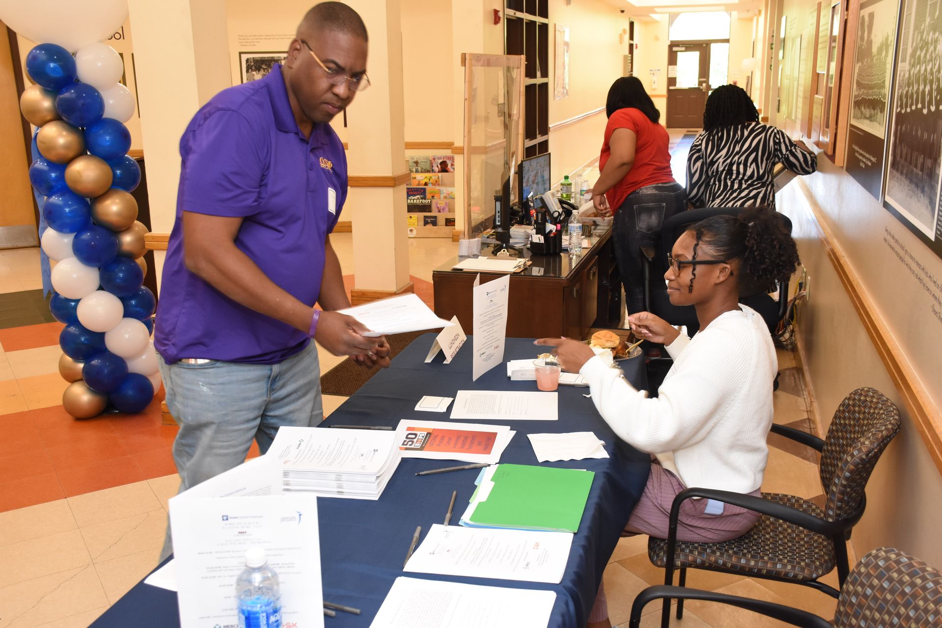 Man at a table with papers interacting with a person. Inside a building, balloons on the left.