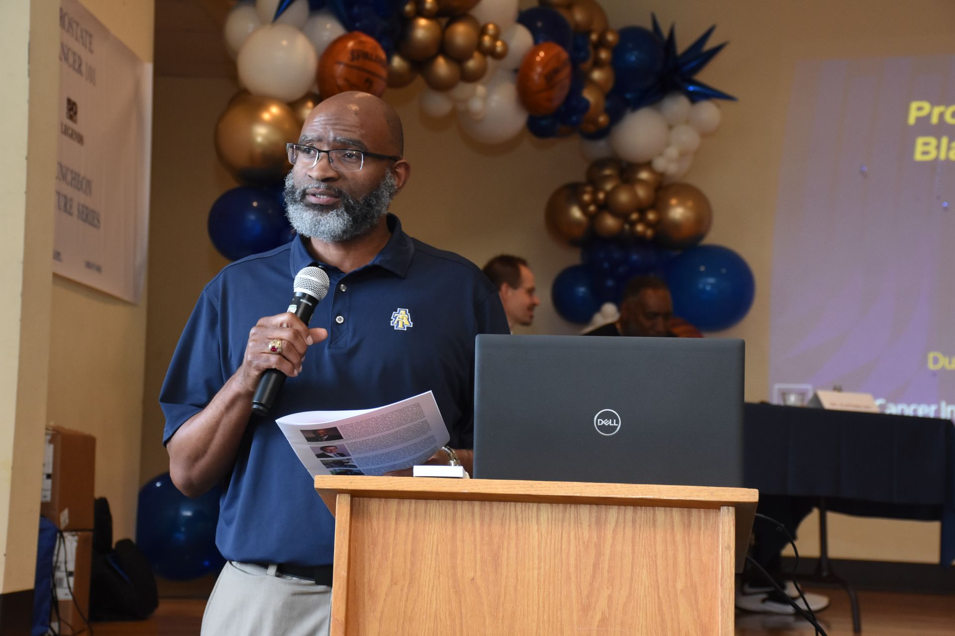 Man speaking at a podium, holding a microphone, with a laptop in front of him. Balloons in the background.
