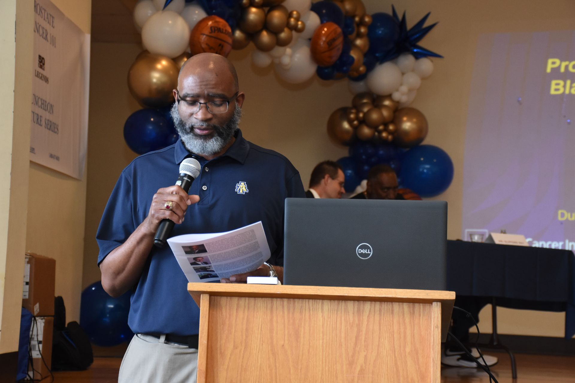 Man speaking at a podium, reading from paper. Laptop, balloons, and another person visible.