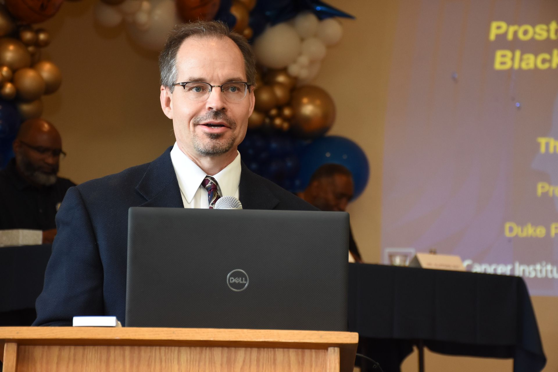 Man at podium with laptop giving a presentation. Balloons and sign in the background.