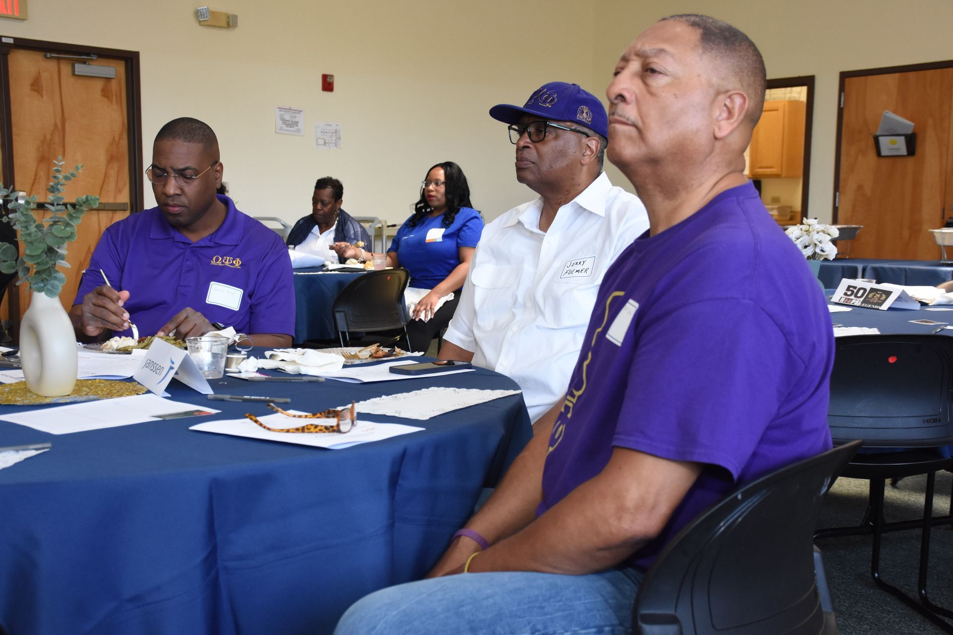 People at a meeting, some wearing purple shirts, seated at tables with blue tablecloths.