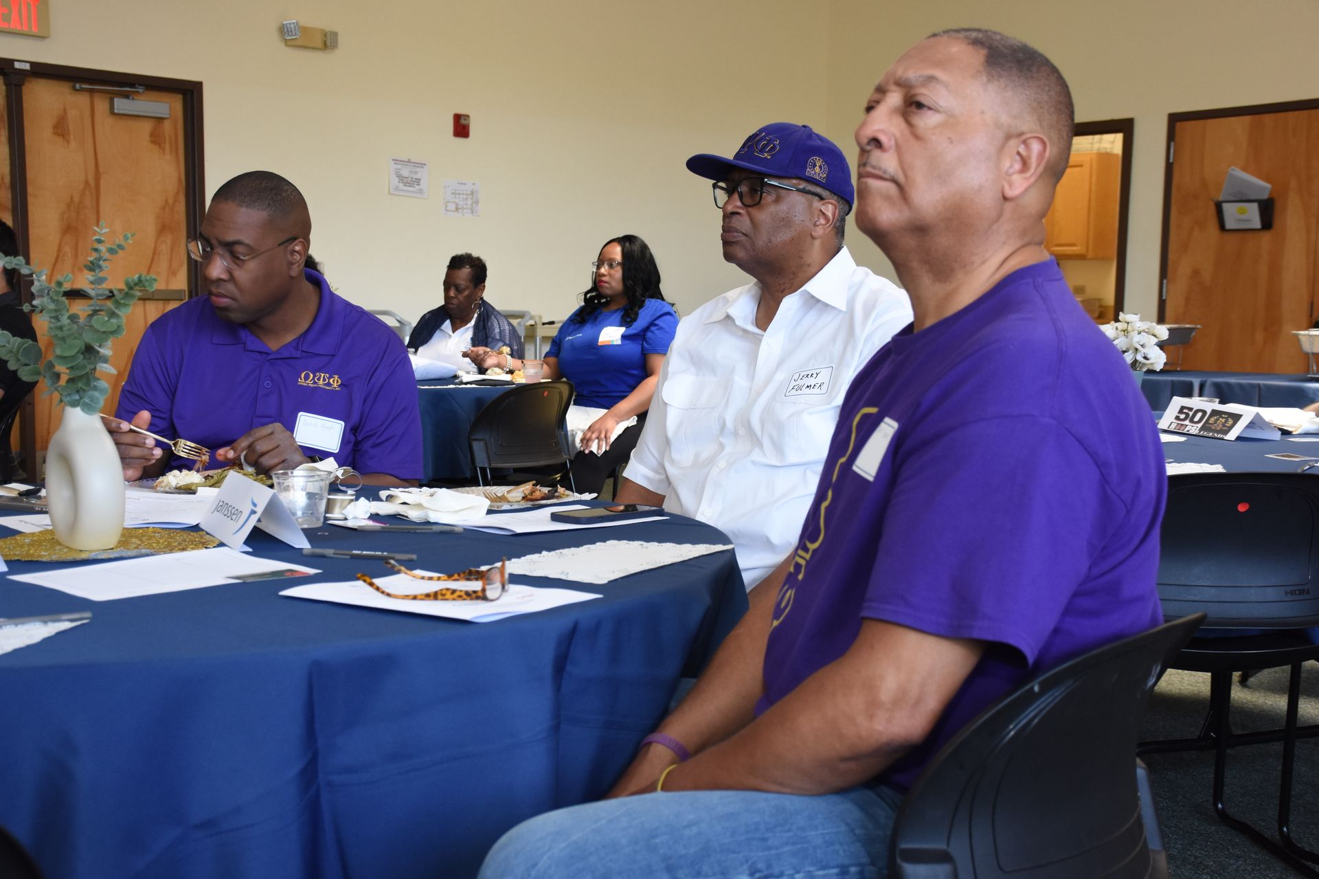 People at a meeting, seated at tables with blue tablecloths. Some are wearing purple and white shirts.