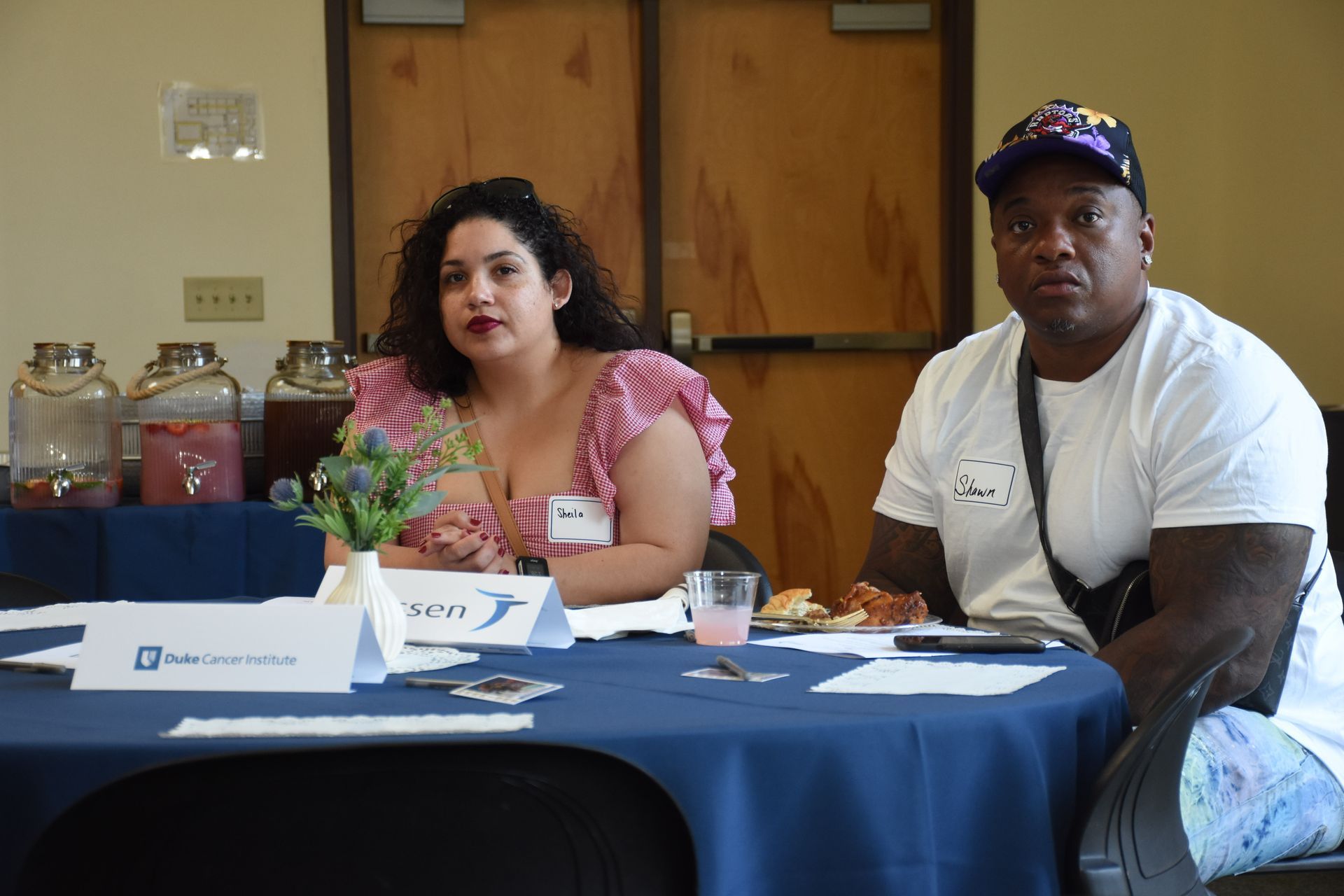 Two people seated at a table with drinks and snacks. Woman in floral top; man in white shirt and cap.