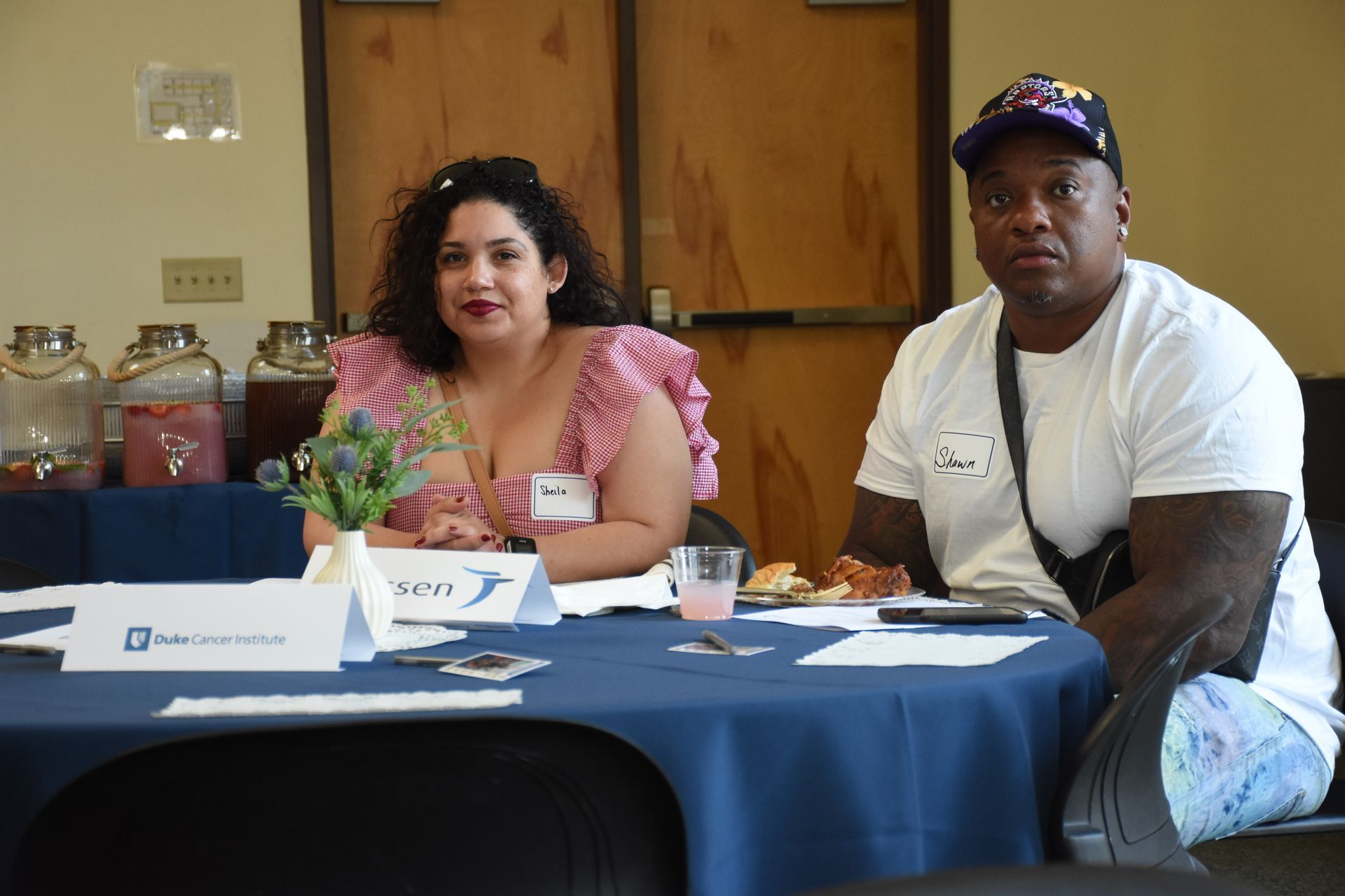 Two people at a table with name tags. Woman with curly hair, pink top. Man in a hat, white shirt.