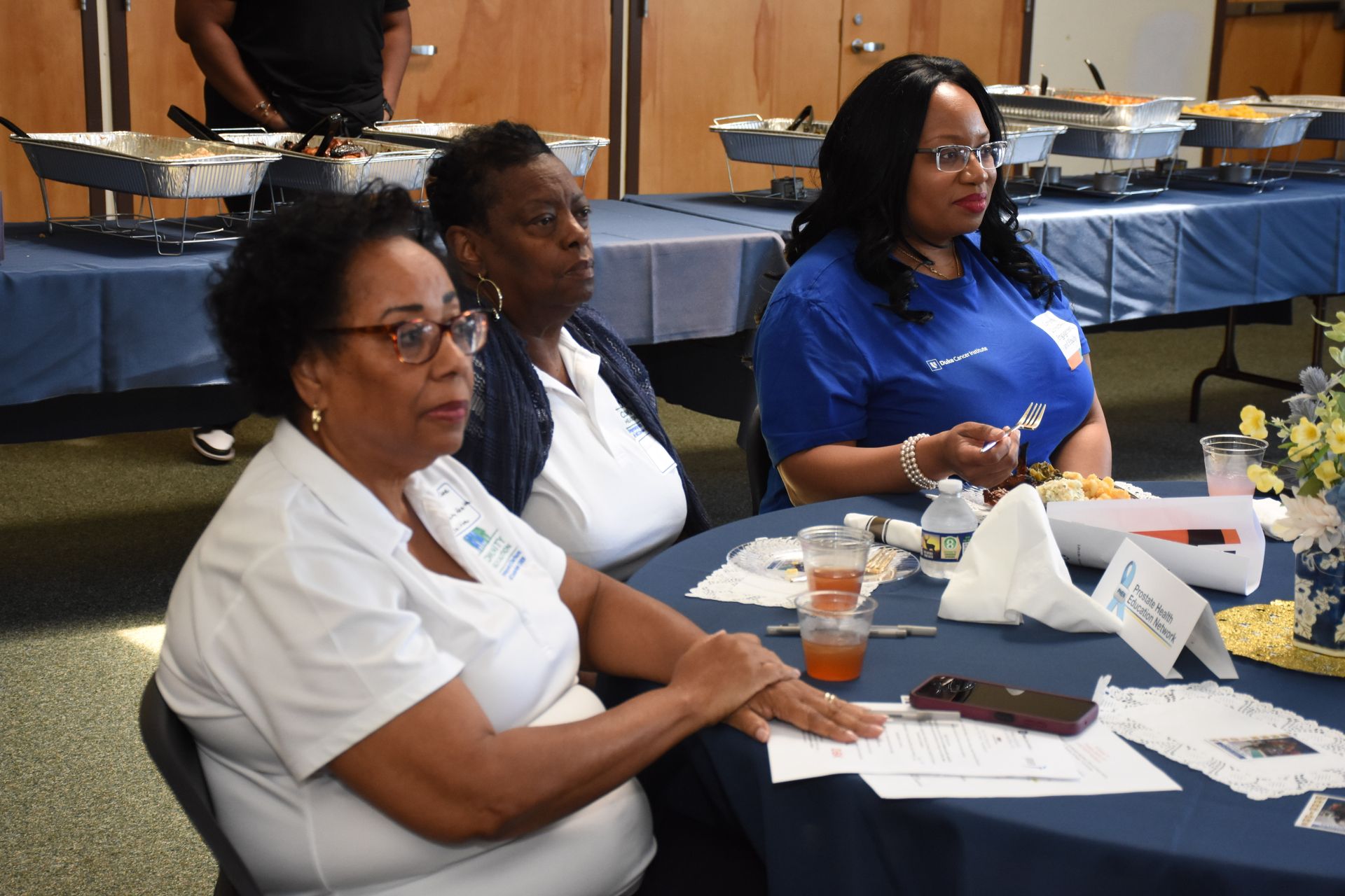 Three women seated at a table with food, listening intently. Dark-haired, wearing glasses, and a blue shirt.