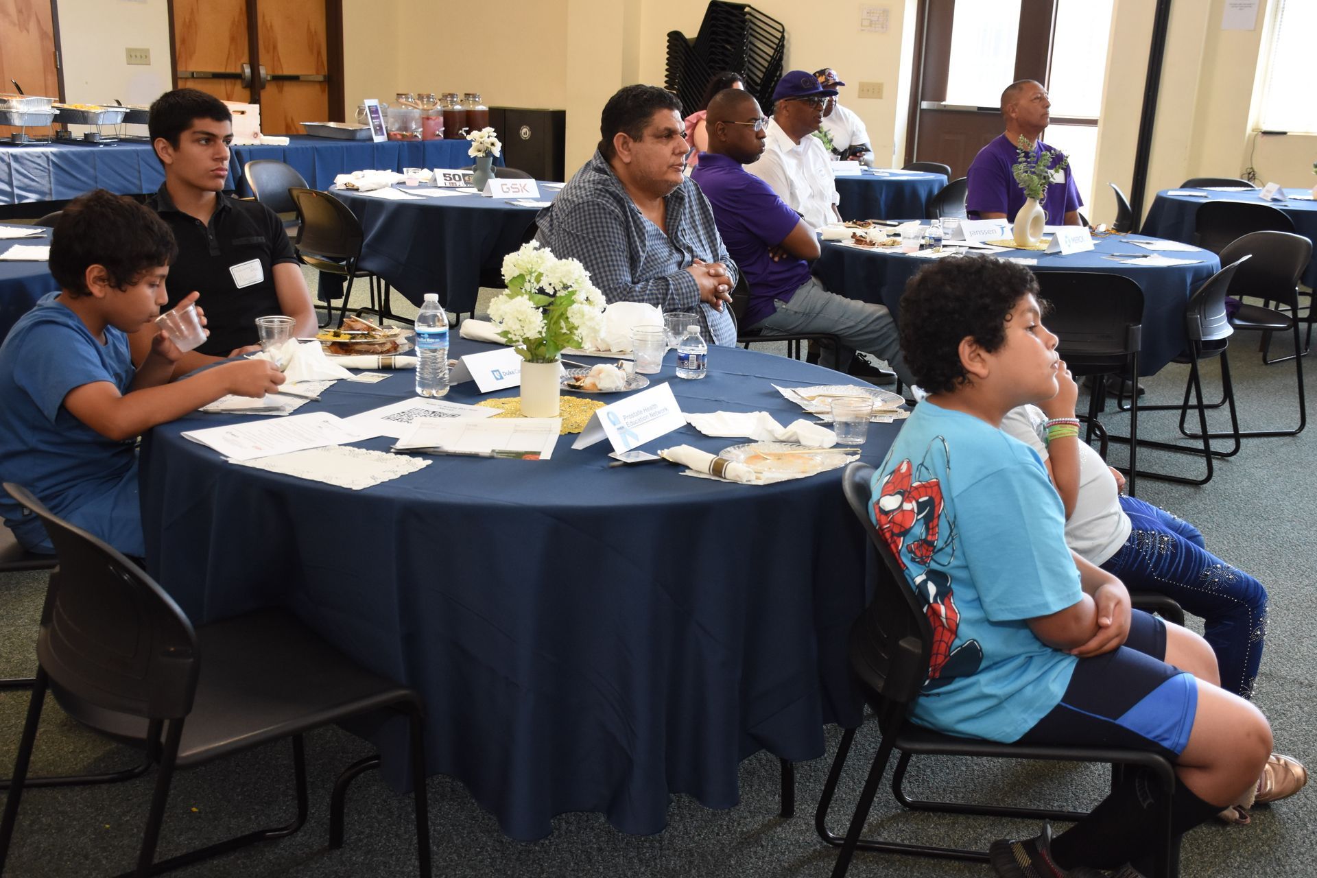 People seated around round tables at an event; blue tablecloths, white flowers, and papers present.