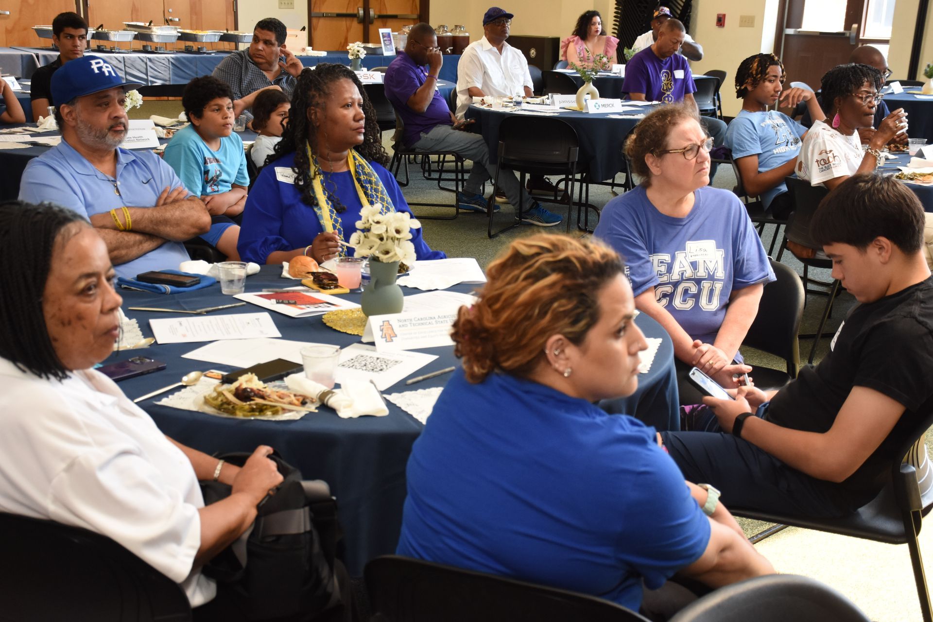 People seated at tables in a room, listening. Some wearing blue shirts, looking attentively.