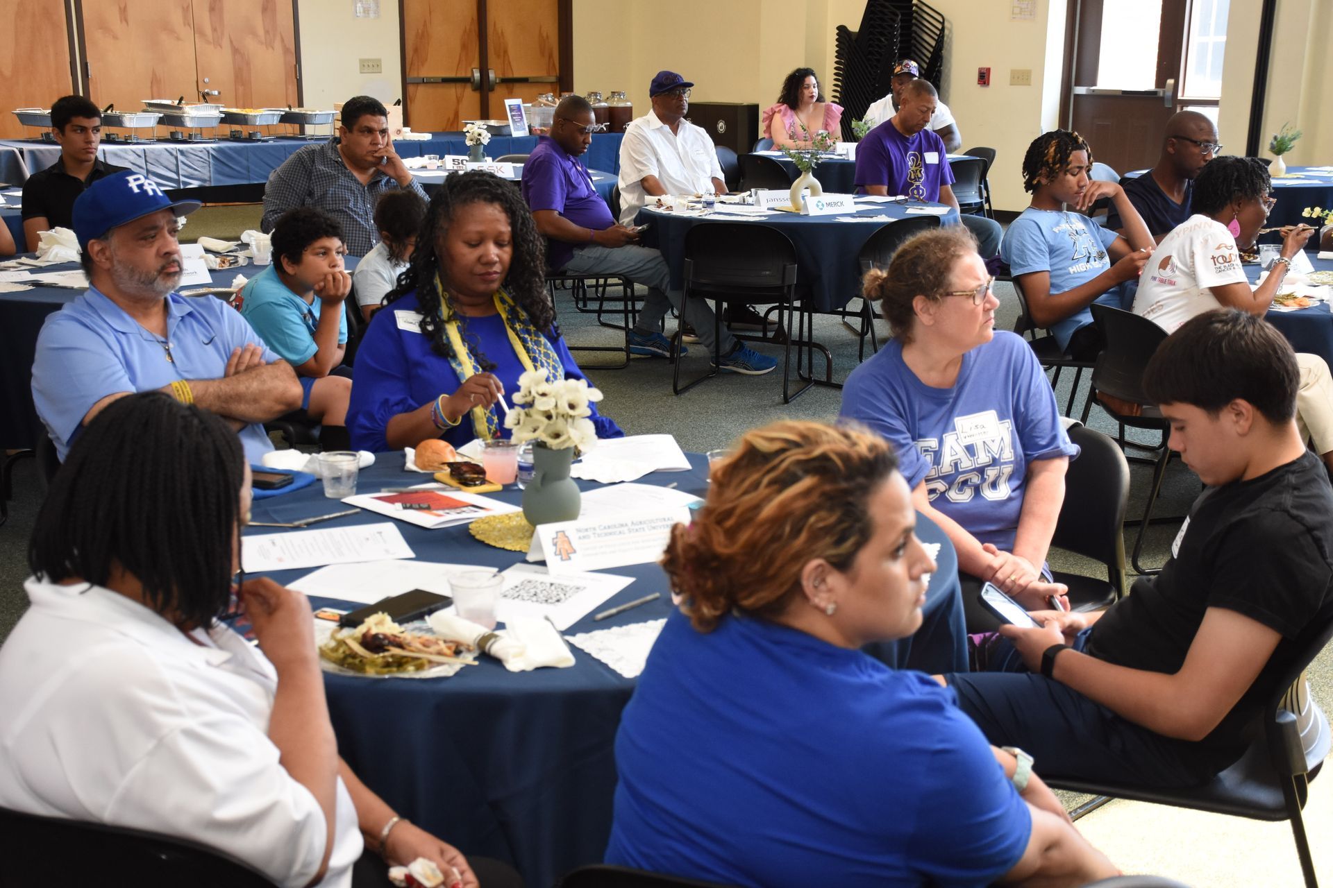 People seated at tables in a room, listening, some wearing blue shirts.