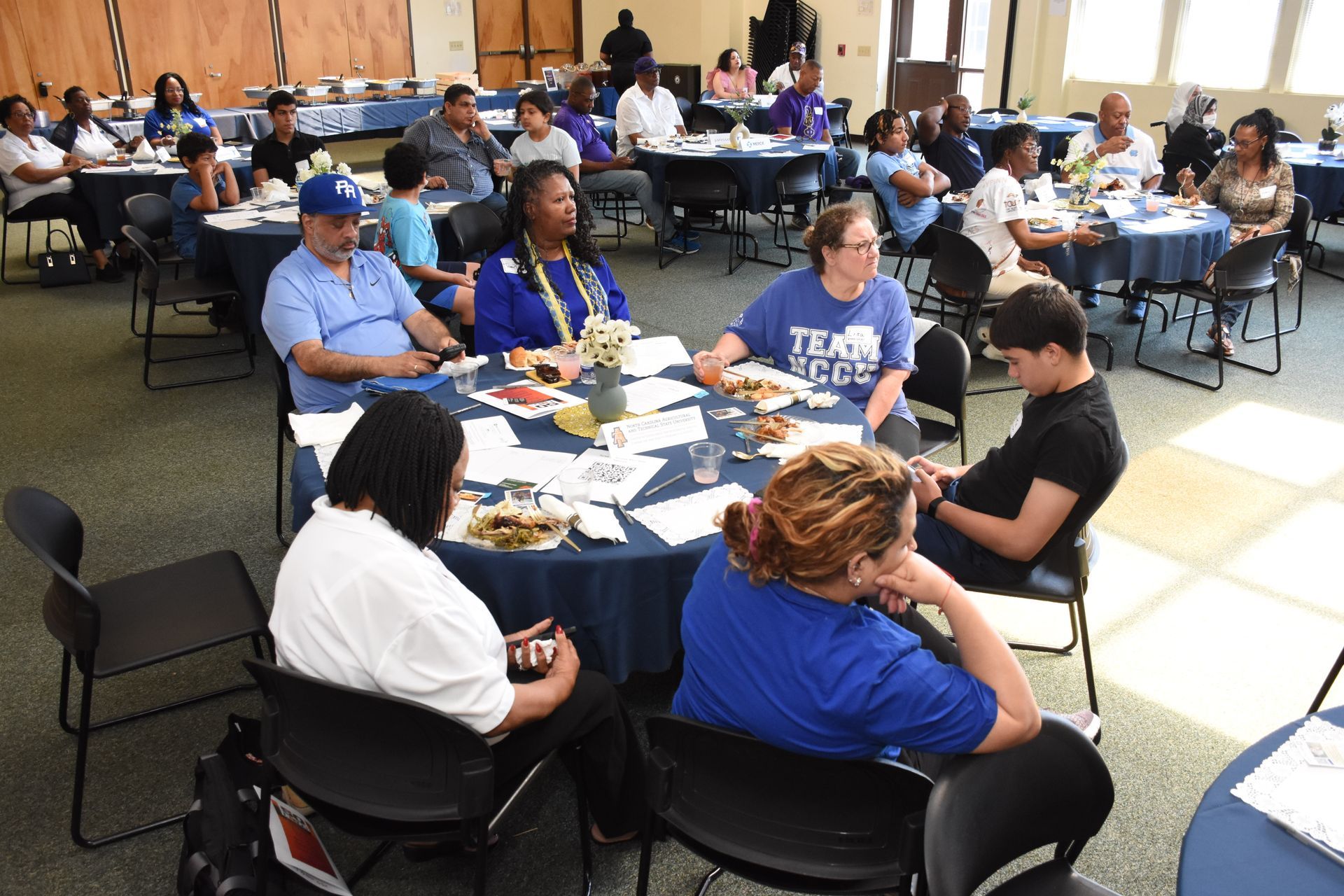 People seated at round tables in a conference room, some wearing blue shirts.