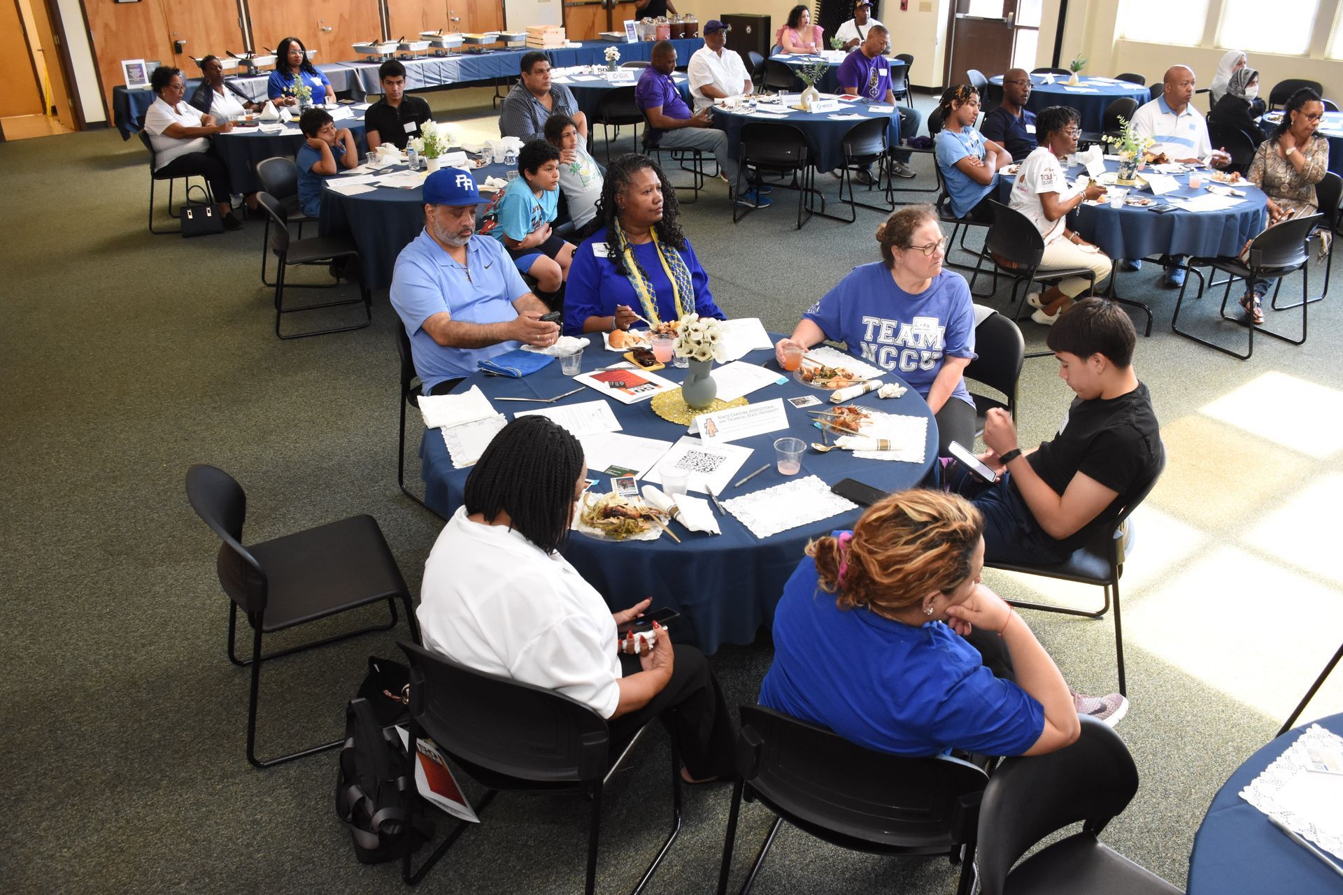 People seated at tables in a large room, attending an event. Tables are covered with blue tablecloths.