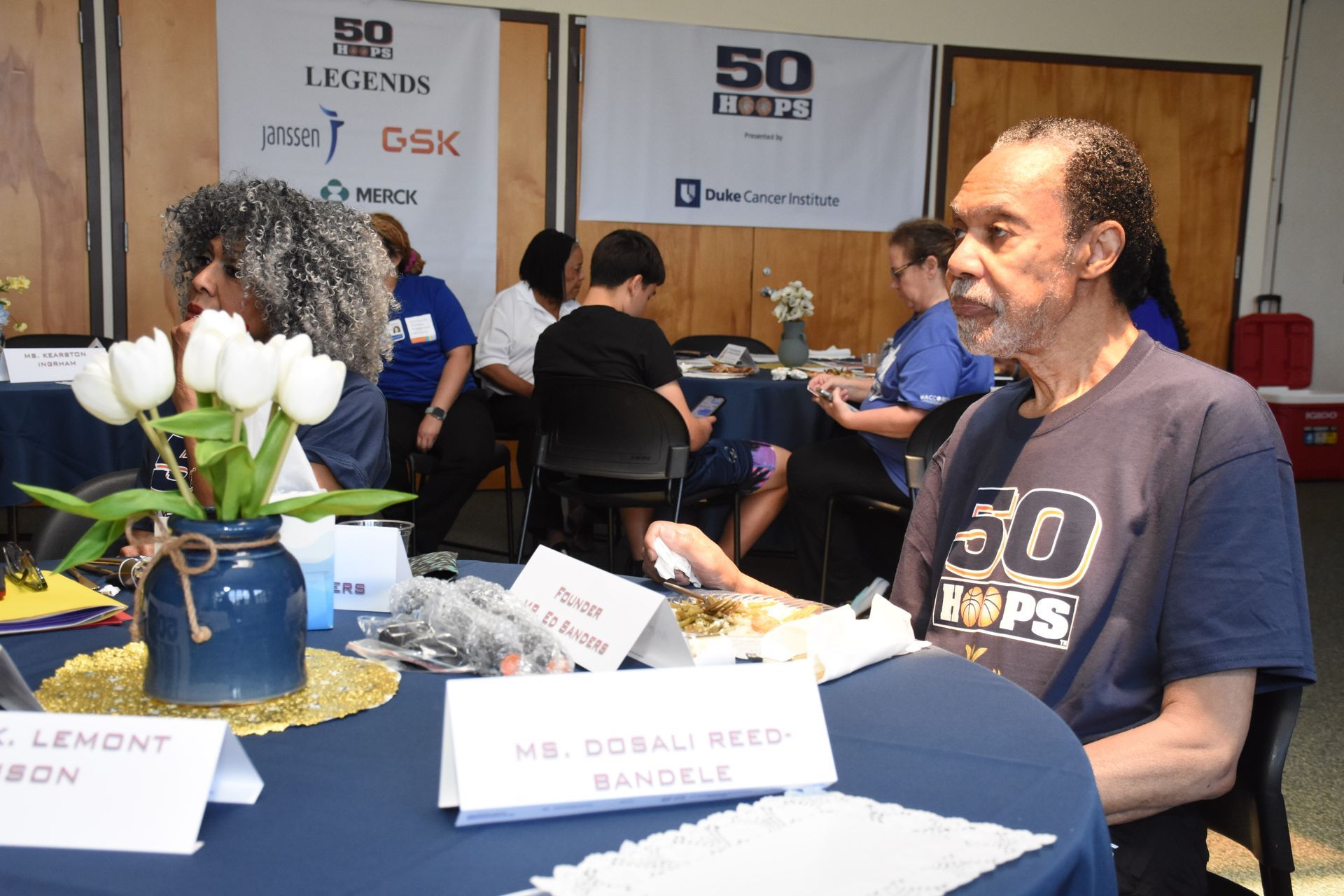 People at an event seated at tables. A man in a blue shirt is in the foreground, with a woman beside him.