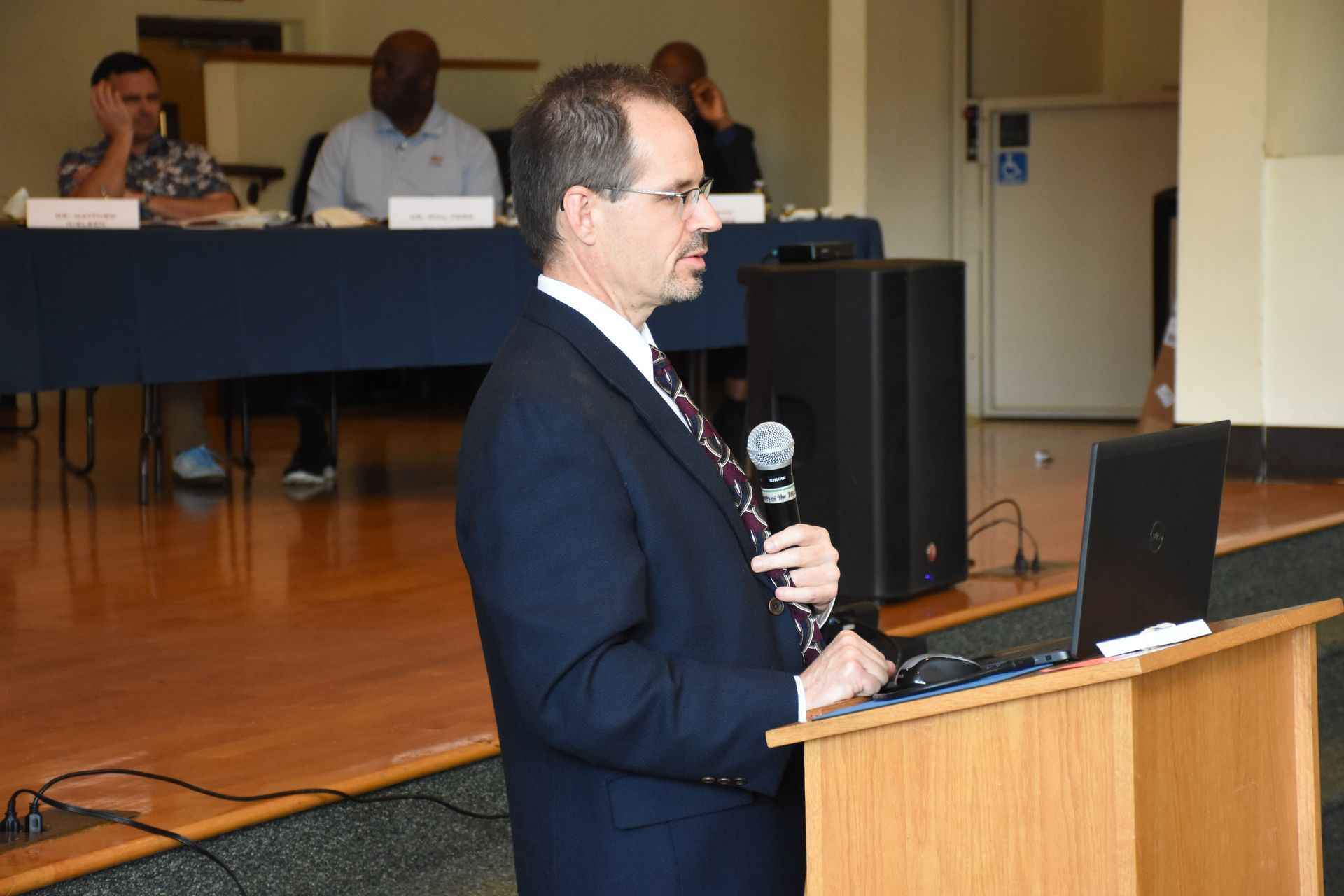 Man in suit speaks at podium, laptop visible. Other people sit at table in background.