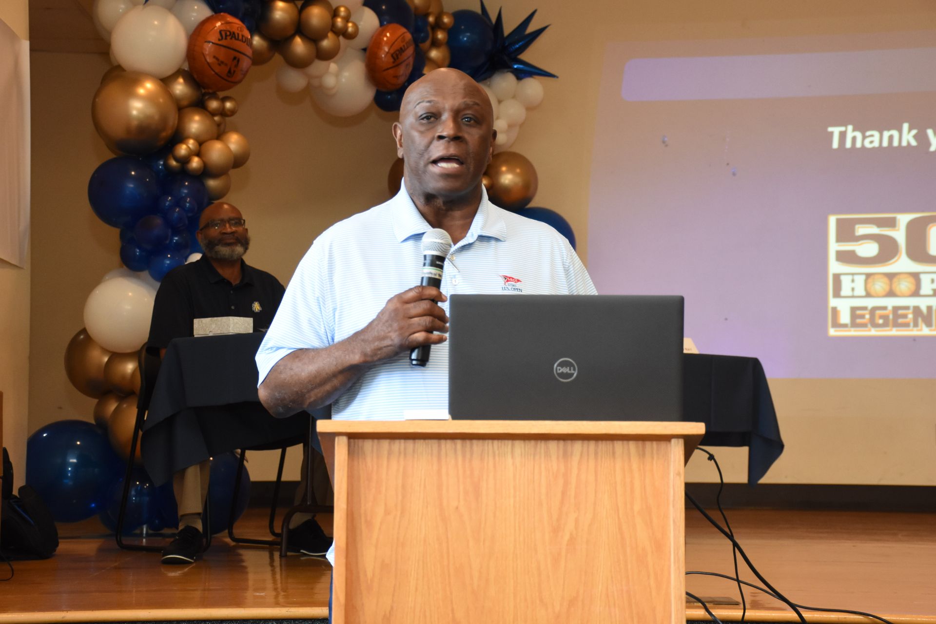 Man speaking at podium, using microphone, with laptop. Balloons in background.