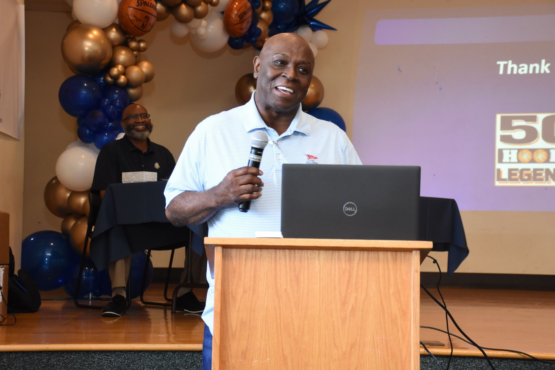 Man speaking at a podium with a laptop, in front of a backdrop with balloons.
