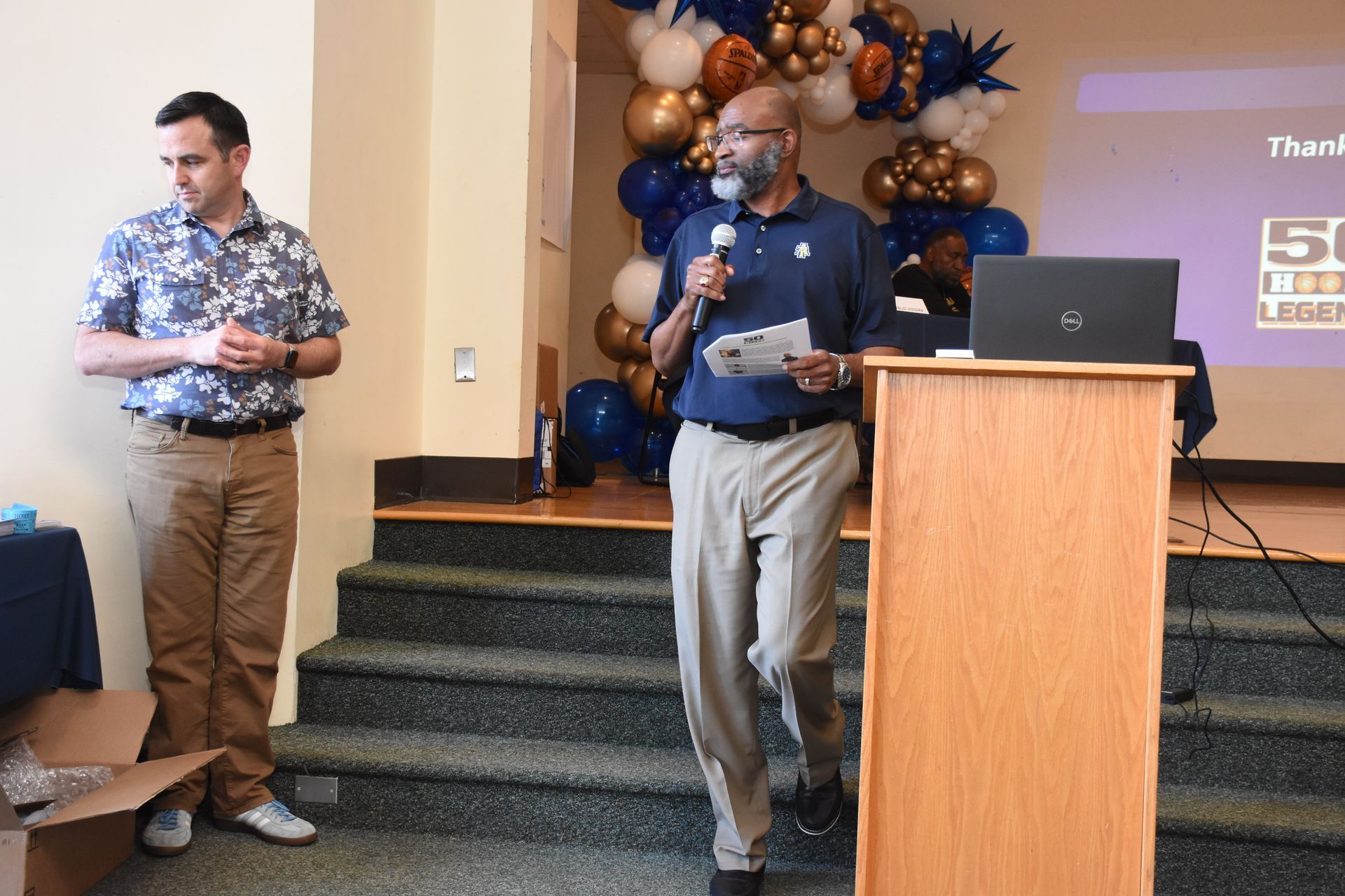 Man speaking at podium, another man beside him, staircase background, balloons.