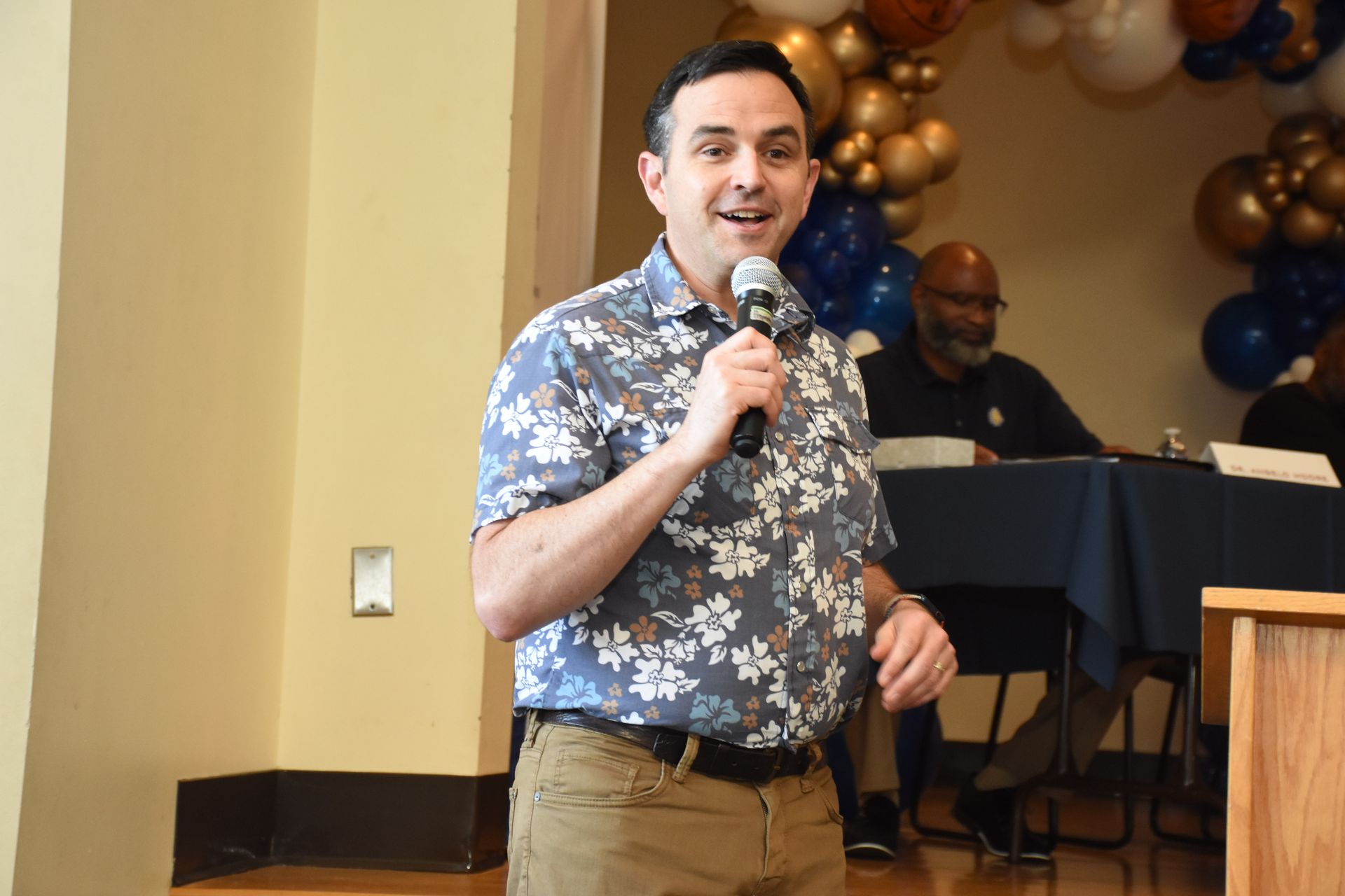 Man speaks into a microphone at a podium, wearing a floral shirt. Balloons decorate the background.