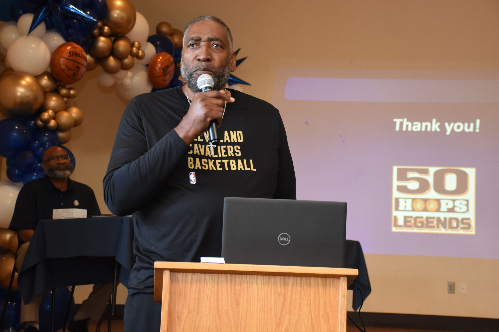 Man speaking at a podium, holding a microphone, with a laptop in front of him. 