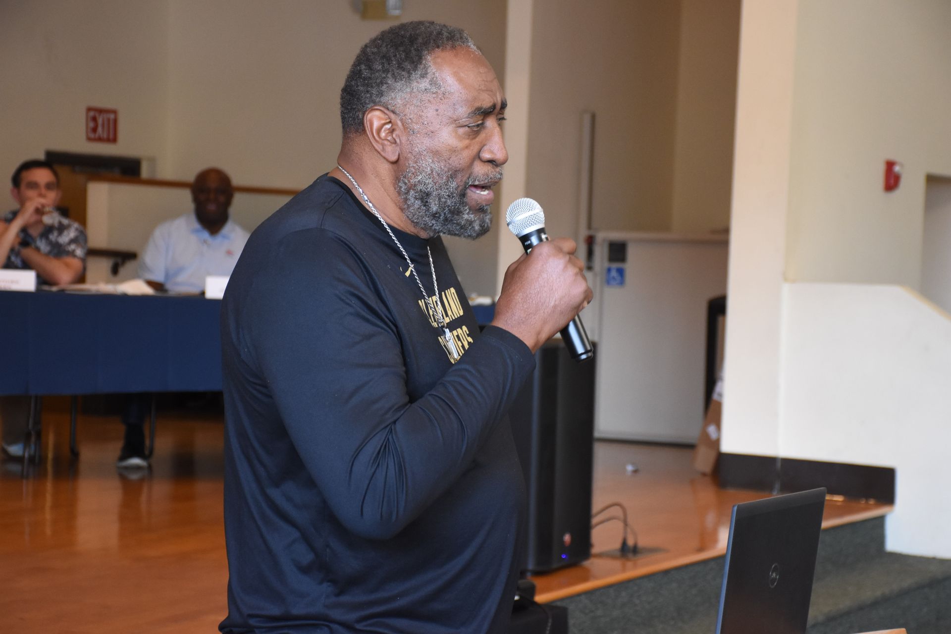 Man speaking into microphone; indoors, wooden floor.