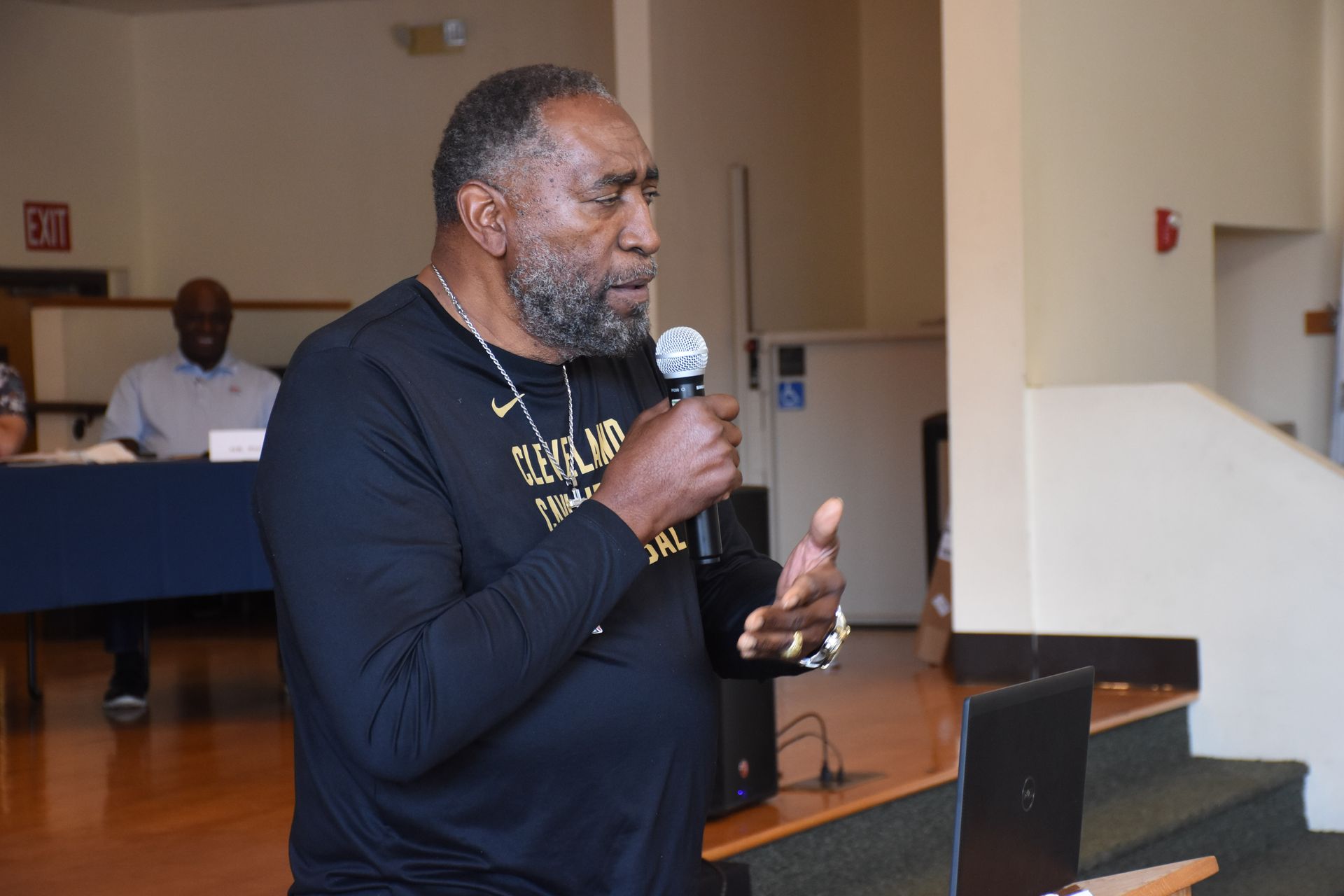 Man speaking into microphone, gesturing with his hands. He's wearing a black shirt, standing at a podium.