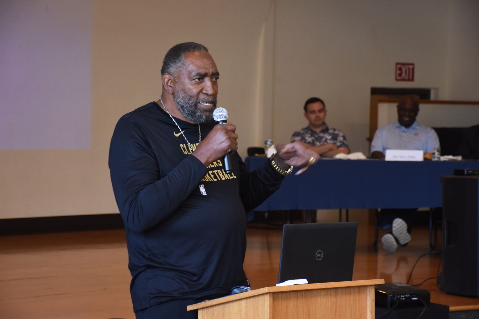 Man speaking at a podium, holding a microphone. Two men sit behind a table in the background.