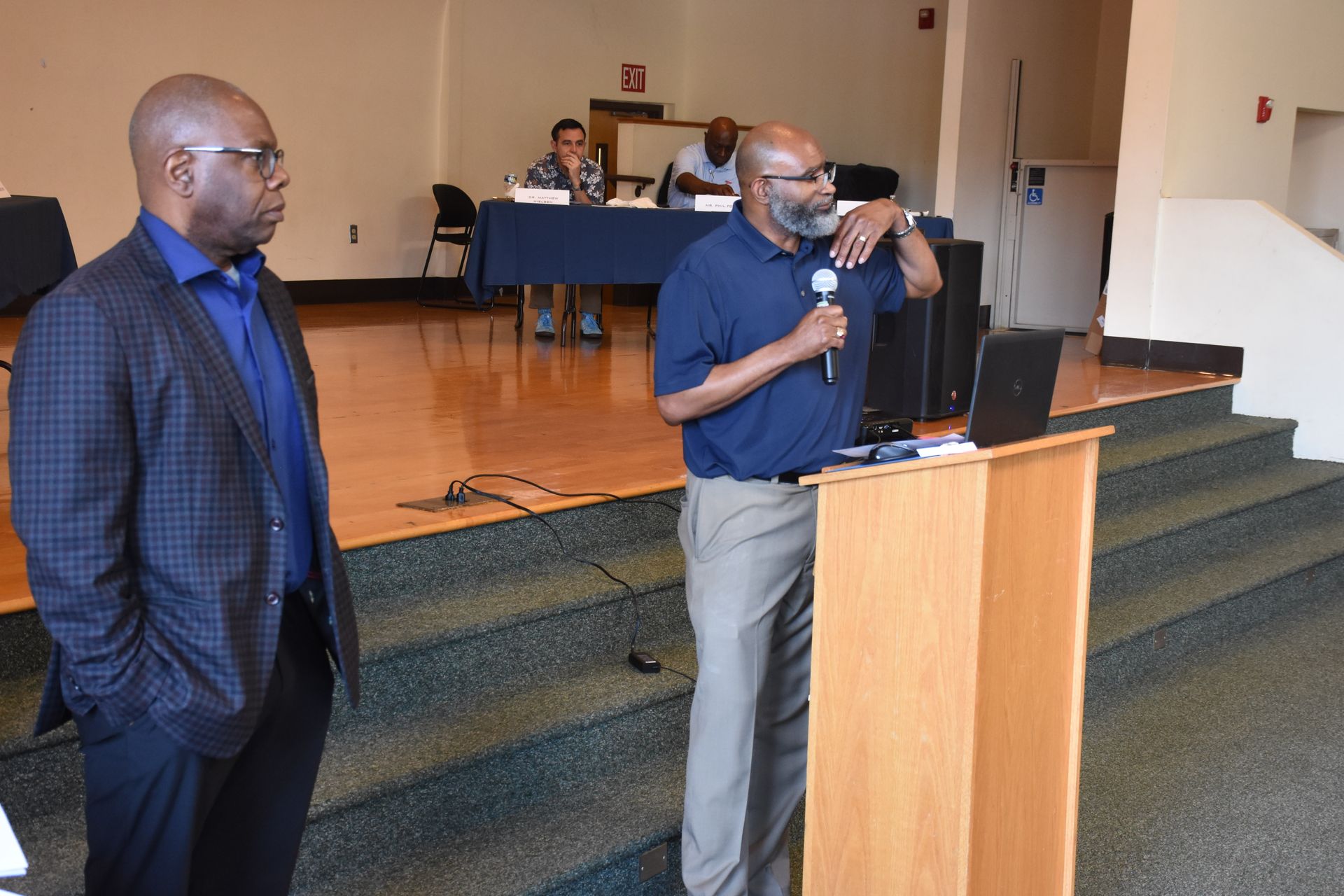 Man speaking at podium, another man stands beside him. Meeting in room with others at a table.