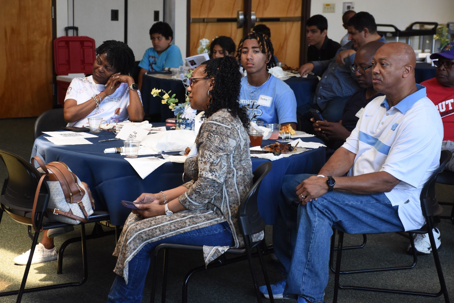 People seated at tables in a room, listening. Some are looking forward, others looking down, with papers and food visible.