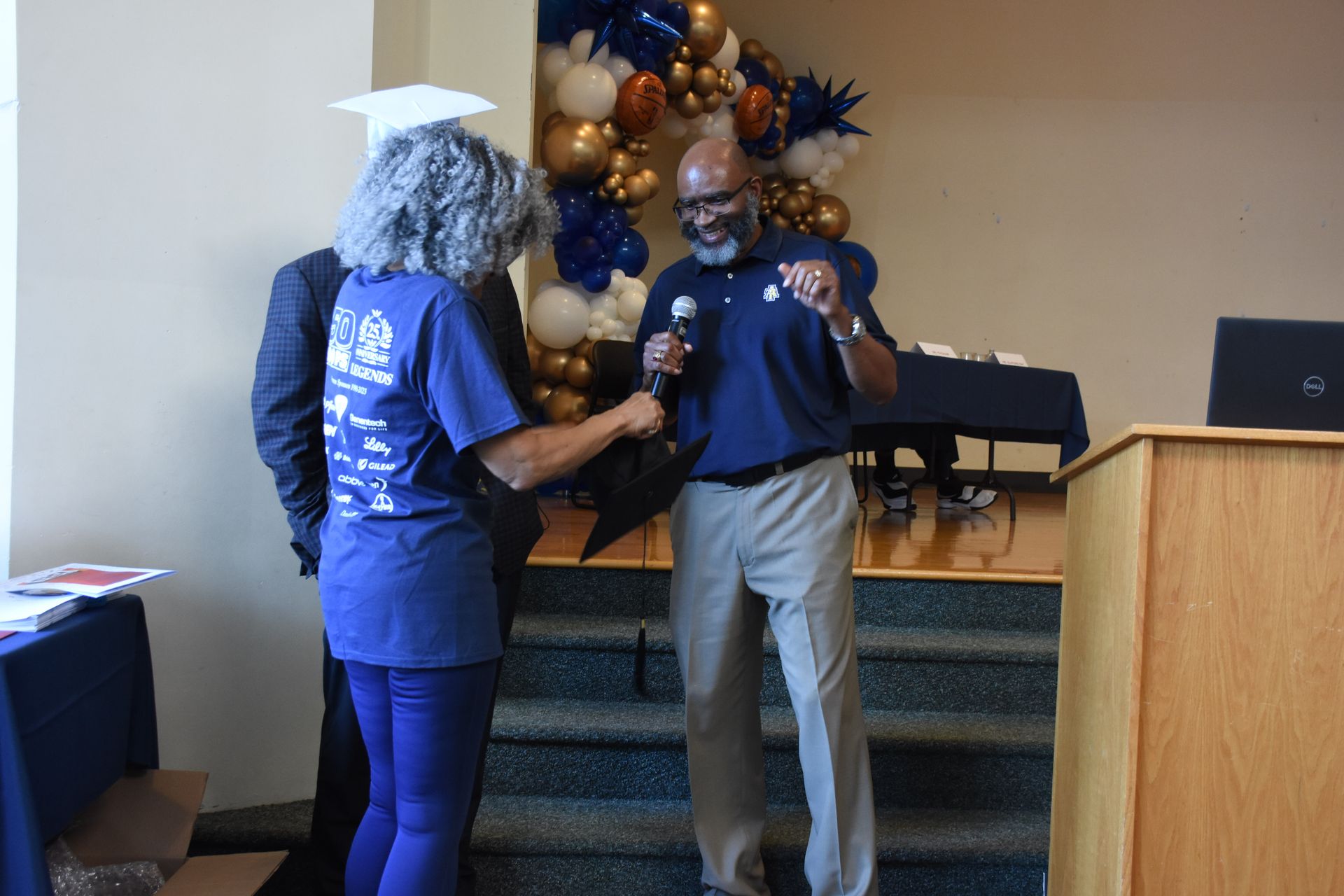Man speaking into microphone; woman adjusting it. Balloons and podium in background.