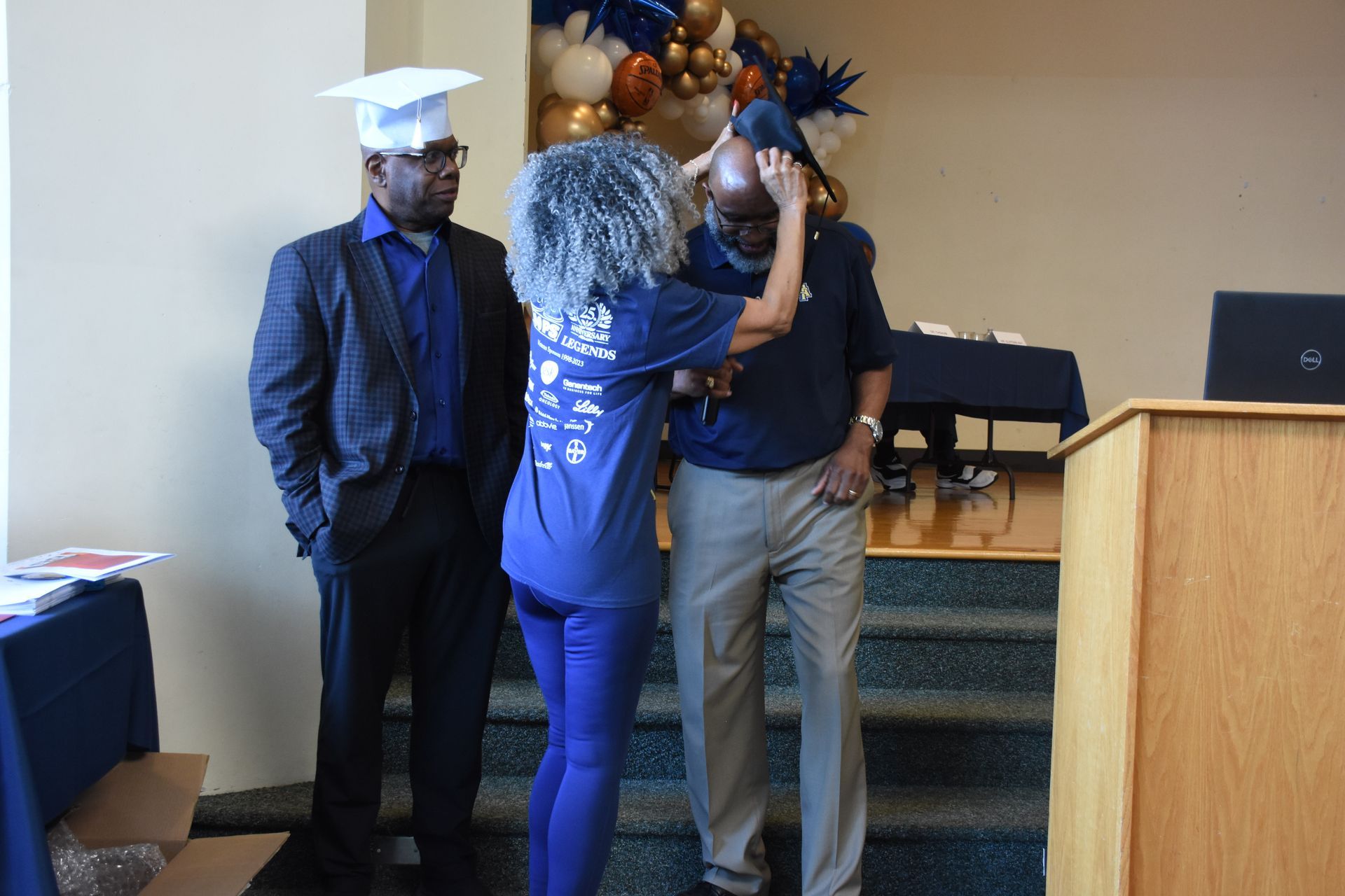 Person being helped with a graduation cap indoors, other person watching. Balloons in the background.